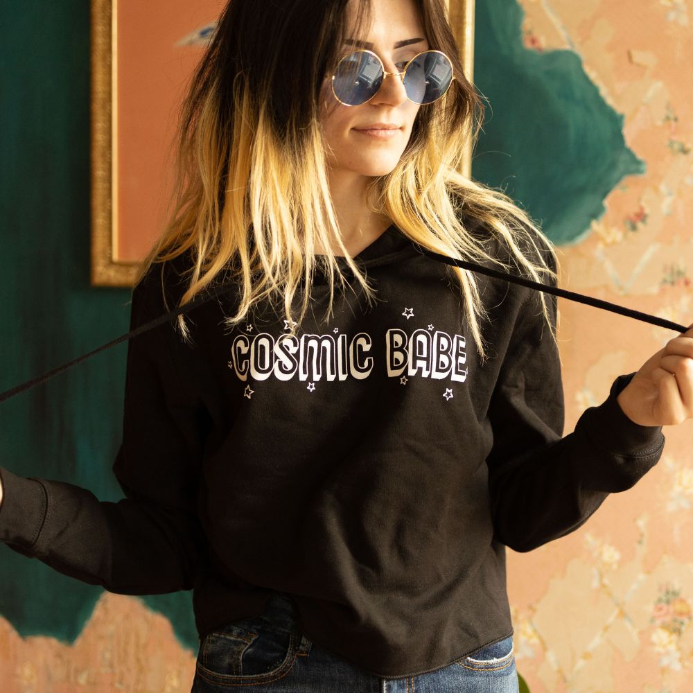The Funnel Cake Tree “Cosmic Babe” cropped raw-edge black hoodie featuring bold white retro lettering with star accents, styled casually with jeans for a cozy celestial look.