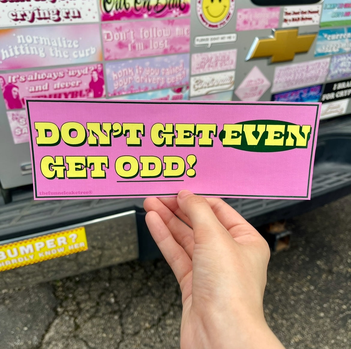 Pink Bumper sticker with yellow text that reads Don't Get Even Get Odd