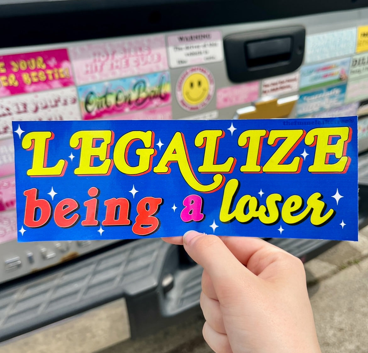 Isabel from The Funnel Cake Tree Holding a bumper sticker with a blue background and yellow, red, and pink text that reads Legalize being a loser