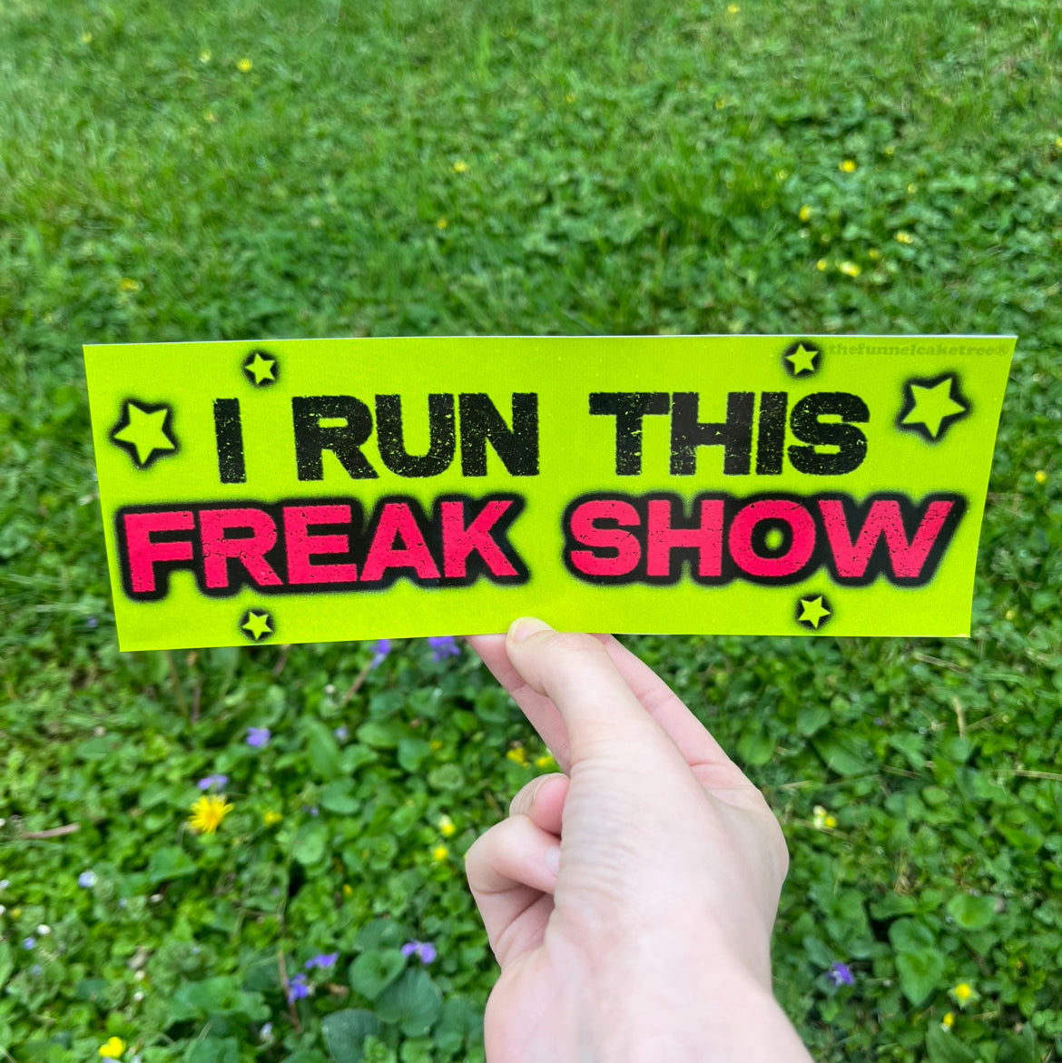 Hand holding green bumper sticker reading I Run This Freak Show
