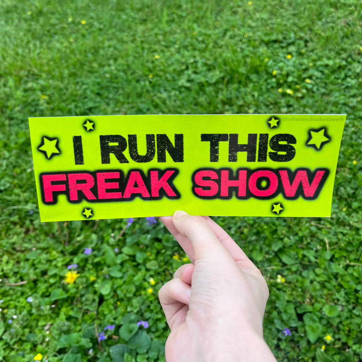 Hand holding green bumper sticker reading I Run This Freak Show