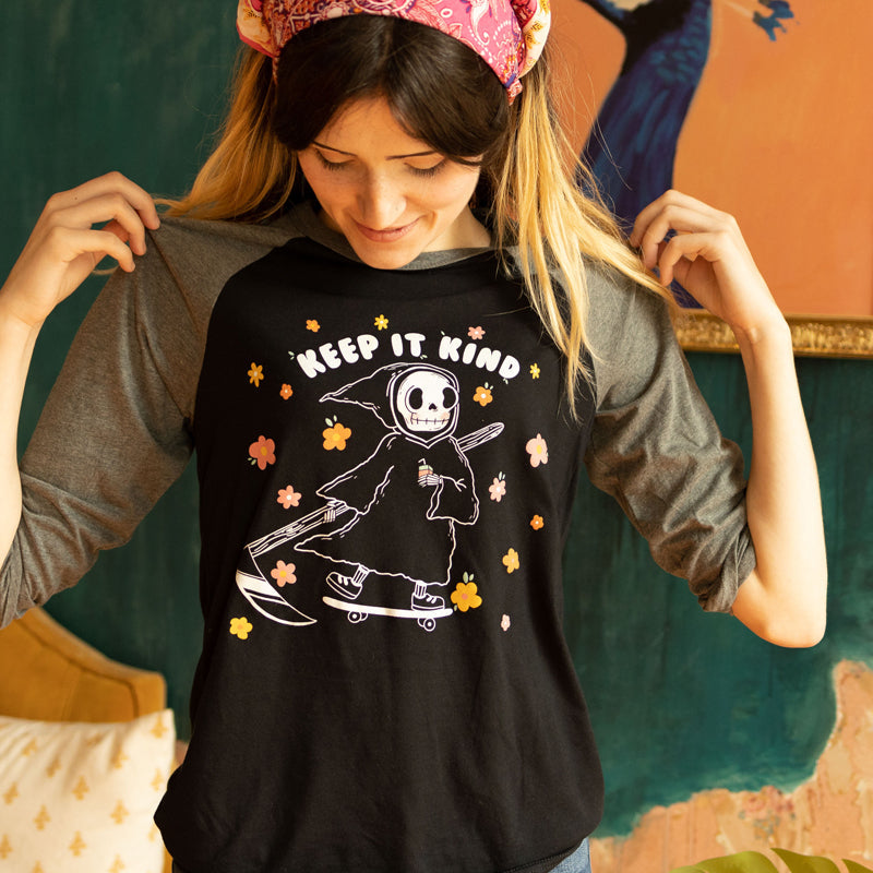 Woman wearing a black and grey raglan tee with a cute cartoon grim reaper holding a coffee and riding a skateboard surrounded by pastel flowers, with the phrase “Keep It Kind” soft retro graphic tee from The Funnel Cake Tree.