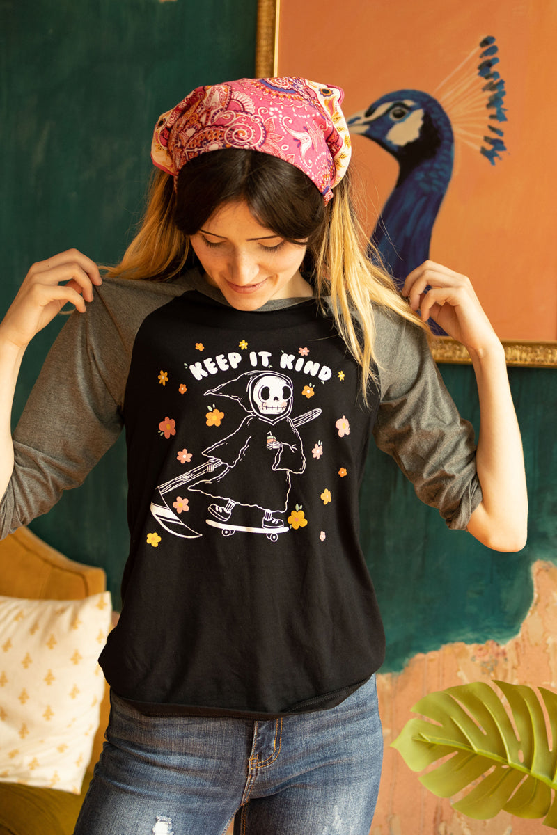 Woman wearing a black and grey raglan tee with a cute cartoon grim reaper holding a coffee cup and riding a skateboard surrounded by colorful flowers, with the text “Keep It Kind” — retro-style graphic tee from The Funnel Cake Tree.
