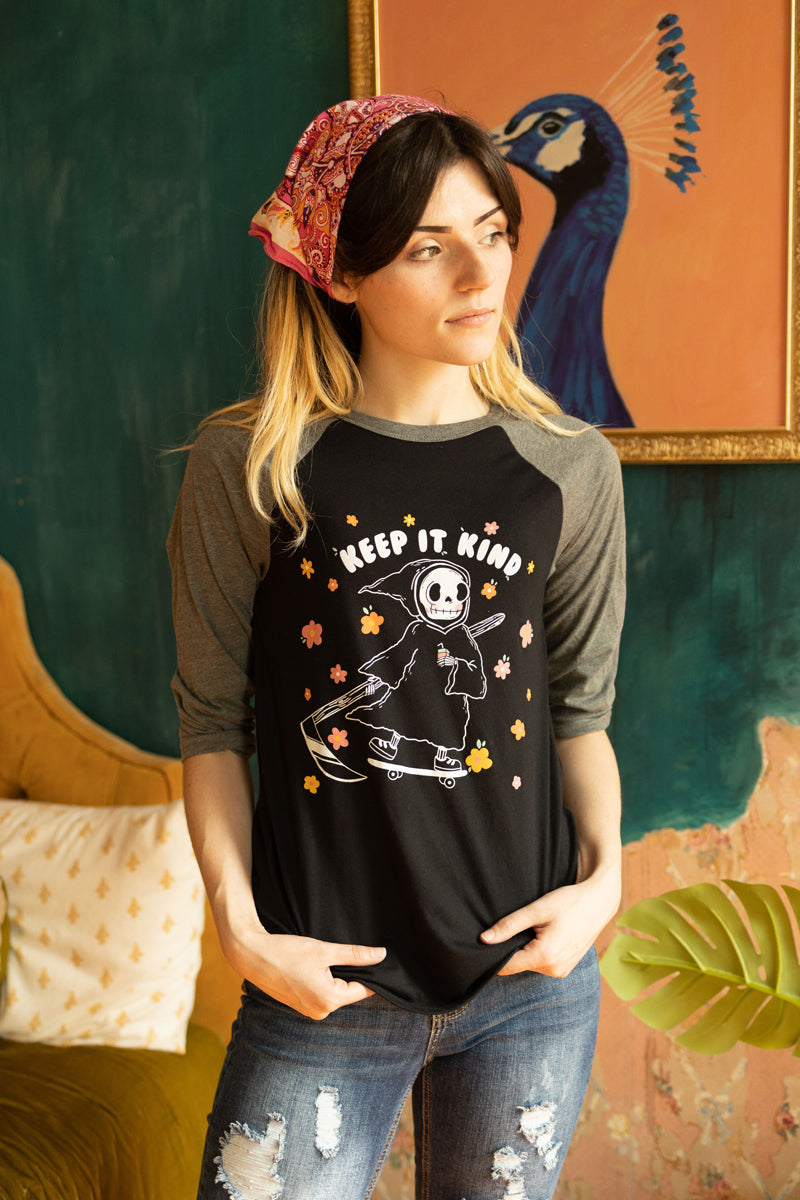 Woman wearing a black and grey raglan tee featuring a cute grim reaper on a skateboard surrounded by pastel flowers with the phrase “Keep It Kind” cozy retro graphic tee from The Funnel Cake Tree.