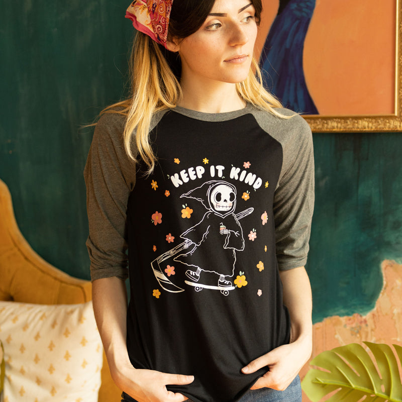 Woman wearing a black and grey raglan tee featuring a cute grim reaper on a skateboard surrounded by pastel flowers with the phrase “Keep It Kind” cozy retro graphic tee from The Funnel Cake Tree.