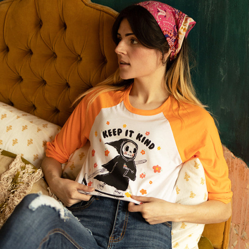 Woman sitting on a vintage mustard couch wearing an orange and white raglan tee that says “Keep It Kind,” featuring a cute cartoon grim reaper on a skateboard surrounded by colorful flowers  retro graphic tee from The Funnel Cake Tree.