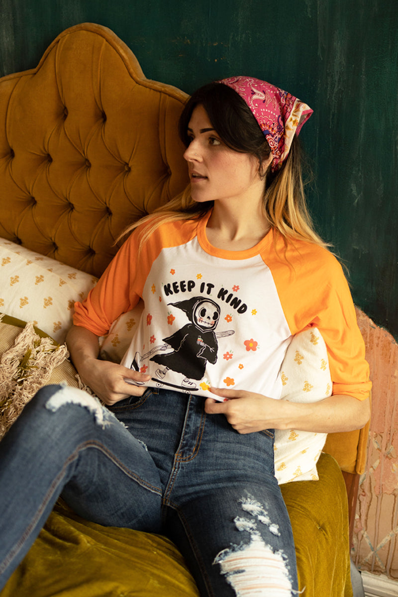 Woman sitting on a vintage mustard couch wearing an orange and white raglan tee that says “Keep It Kind,” featuring a cute cartoon grim reaper on a skateboard surrounded by colorful flowers — retro graphic tee from The Funnel Cake Tree.