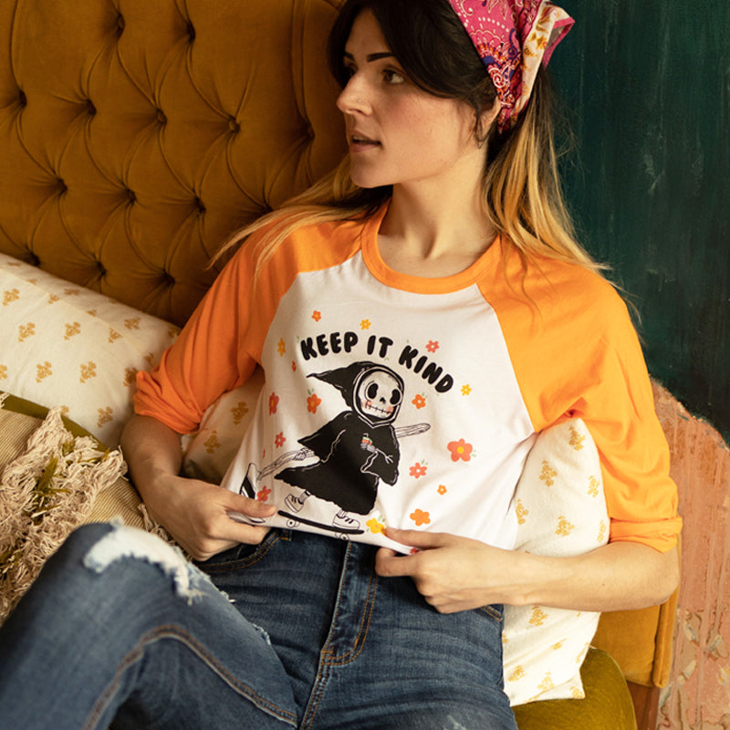Woman sitting on a vintage mustard couch wearing an orange and white raglan tee that says “Keep It Kind,” featuring a cute cartoon grim reaper on a skateboard surrounded by colorful flowers — retro graphic tee from The Funnel Cake Tree.