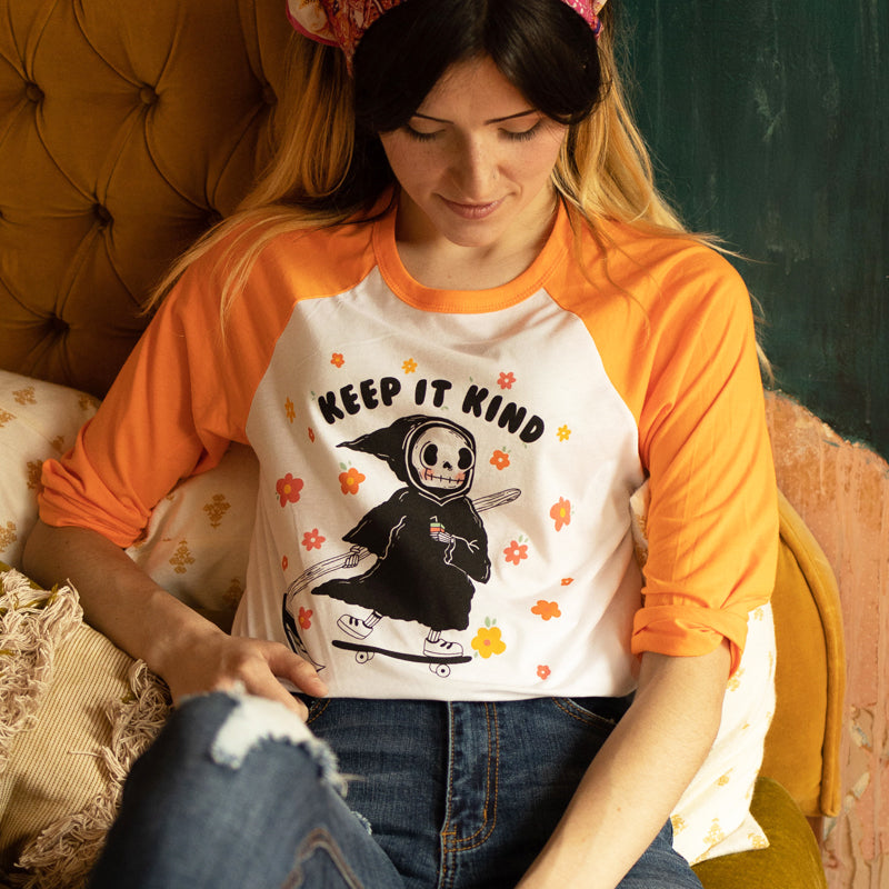 Woman sitting on a vintage mustard couch wearing an orange and white raglan tee with a cute grim reaper holding a rainbow drink and the words “Keep It Kind,” surrounded by cheerful flowers — The Funnel Cake Tree retro graphic tee.