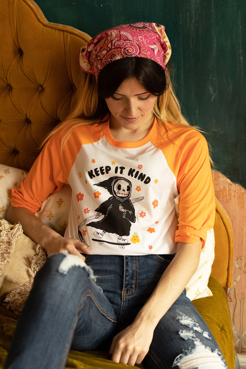 Woman sitting on a vintage mustard couch wearing an orange and white raglan tee with a cute grim reaper holding a rainbow drink and the words “Keep It Kind,” surrounded by cheerful flowers — The Funnel Cake Tree retro graphic tee.