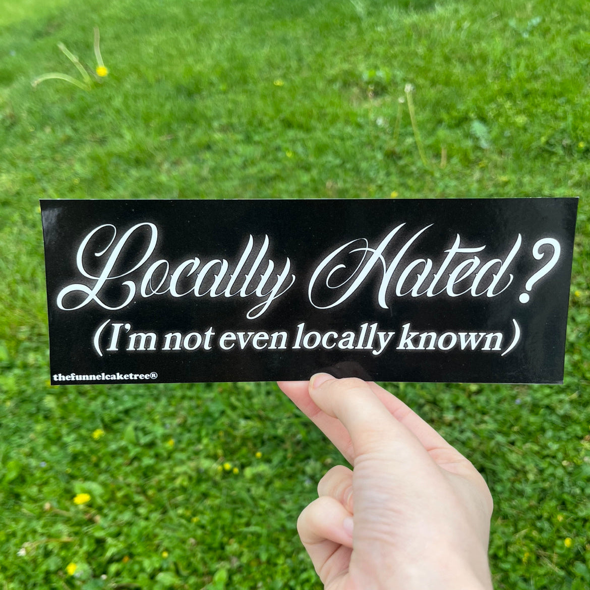 Hand holding black bumper sticker reading Locally Hated I'm not even locally known