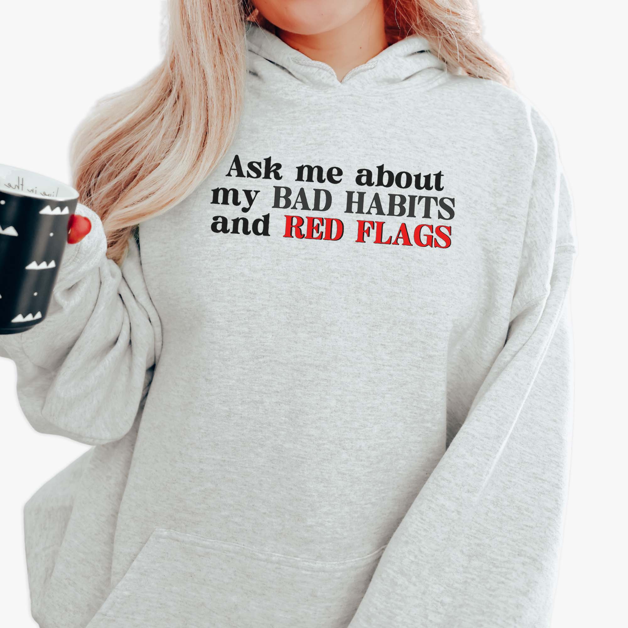Woman wearing a light grey hoodie with black and red text that says “Ask me about my bad habits and red flags” — Funnel Cake Tree pullover hoodie.