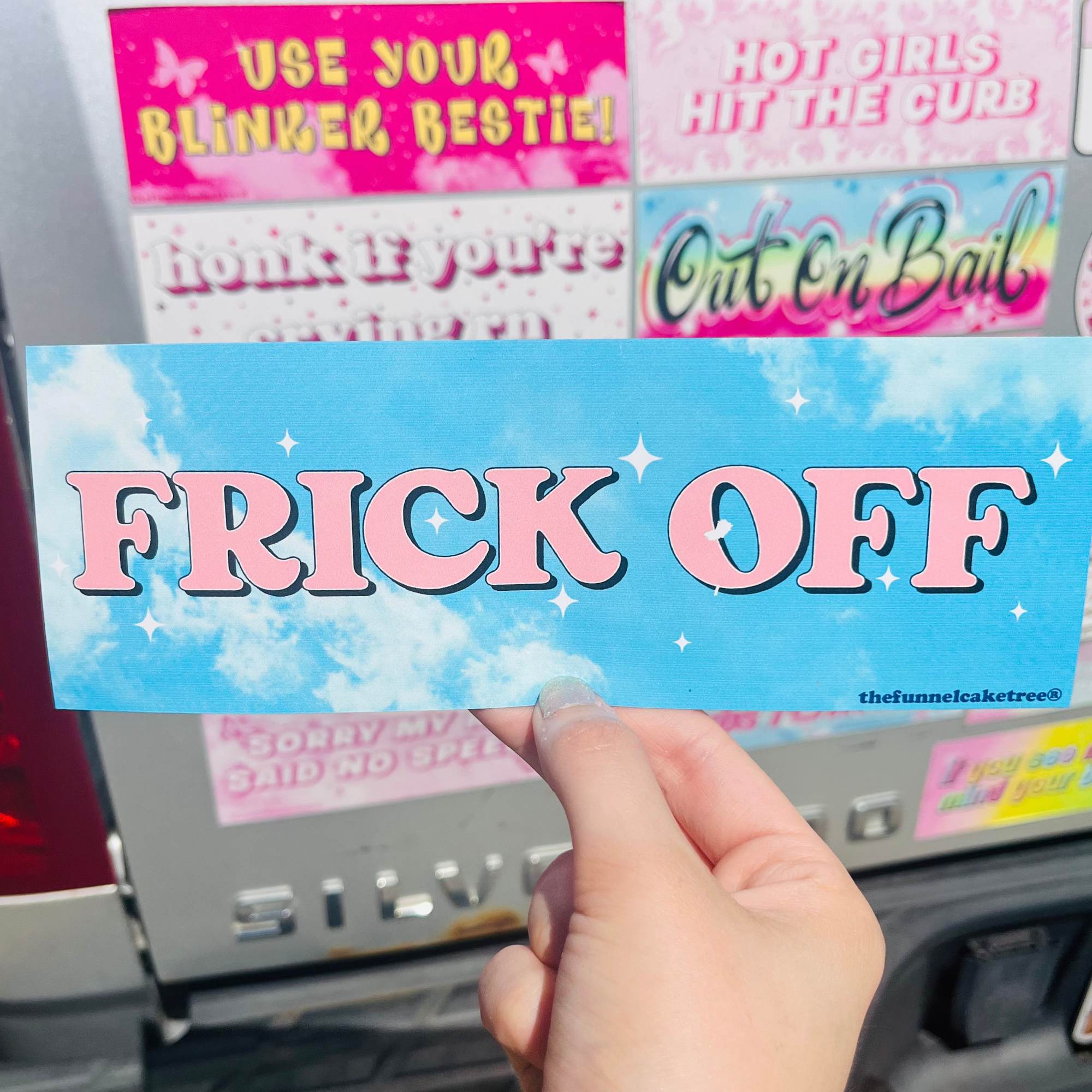 Hand holding a blue bumper sticker with pink text 'FRICK OFF' on a car. Waterproof and UV-protected, perfect for cars