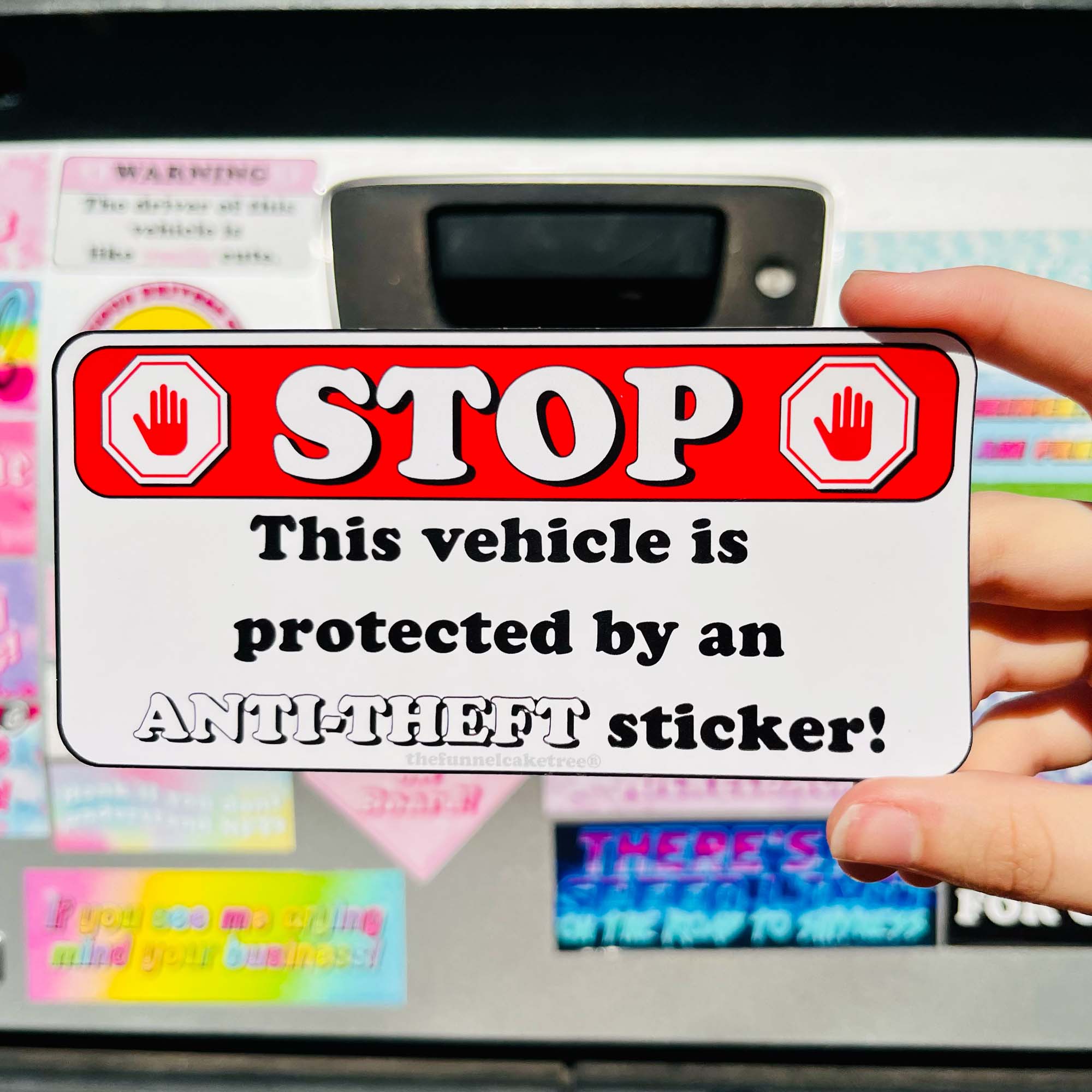 Hand holding a funny and snarky vehicle anti-theft sticker with a red border and white text in front of the back of a car covered in bumper stickers