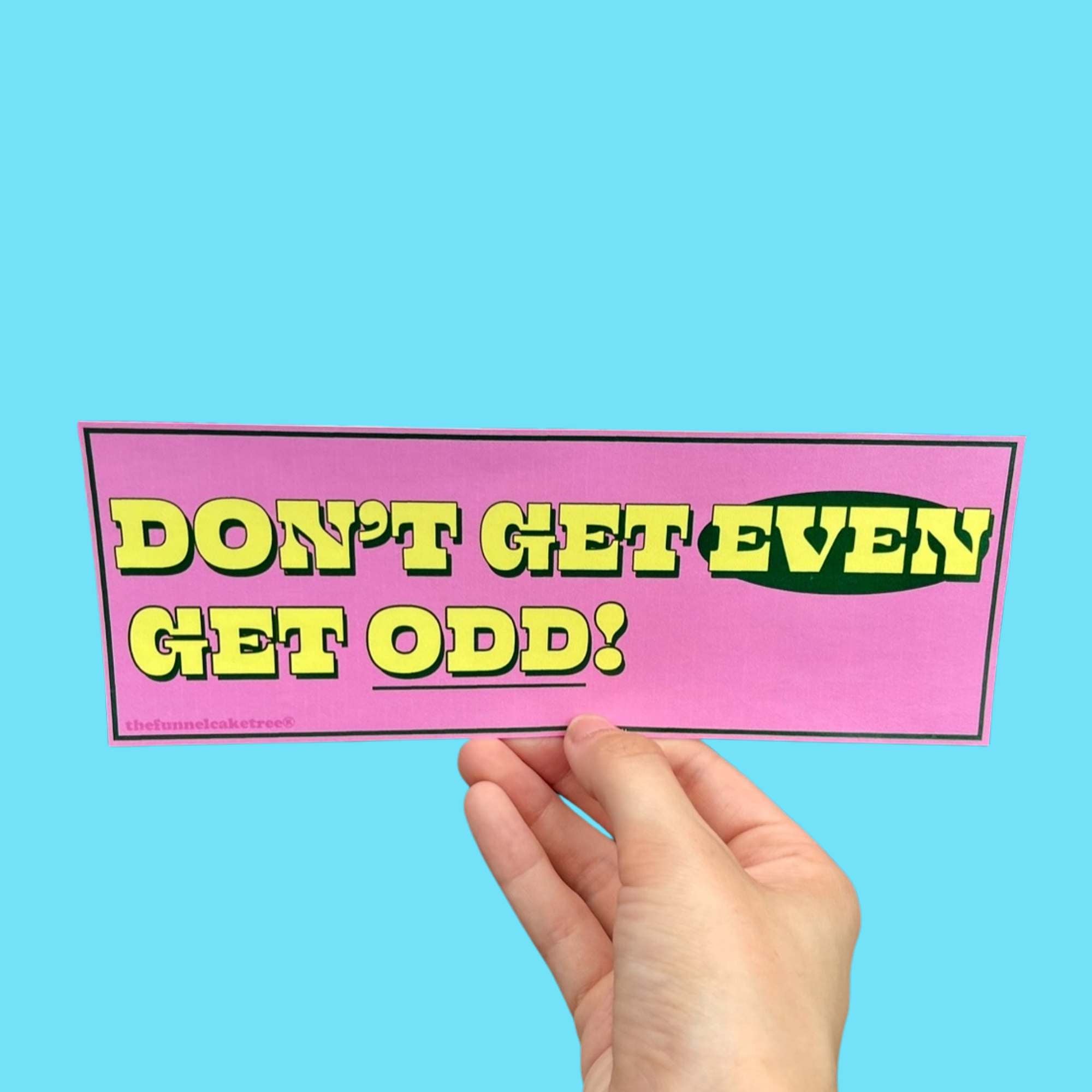 Glossy pink bumper sticker with bold yellow and green text reading “DON’T GET EVEN GET ODD!” held against a light blue background.