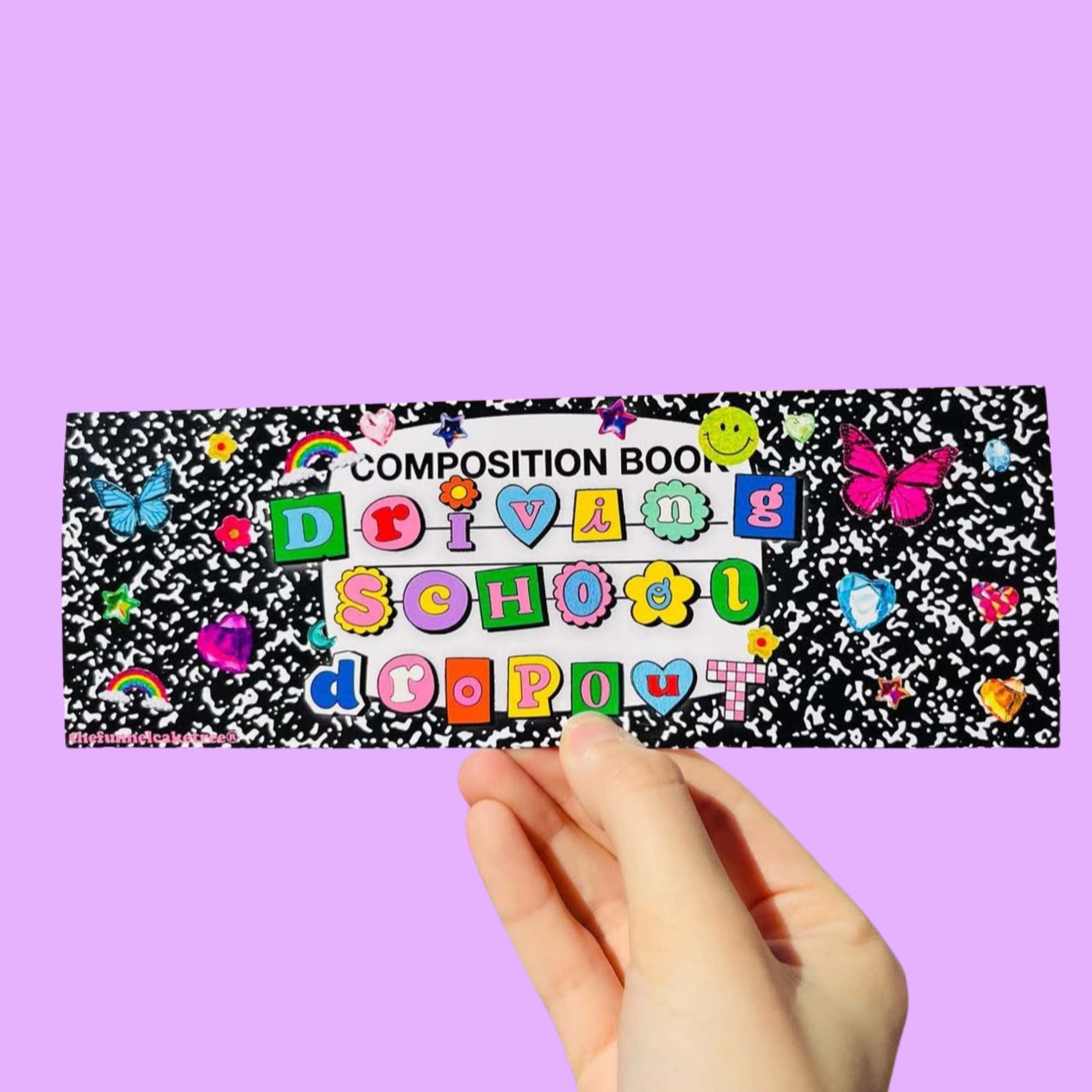  Glossy bumper sticker with black-and-white composition book background and colorful ransom-note letters reading “Driving School Dropout,” decorated with rainbows, butterflies, and smiley doodles.