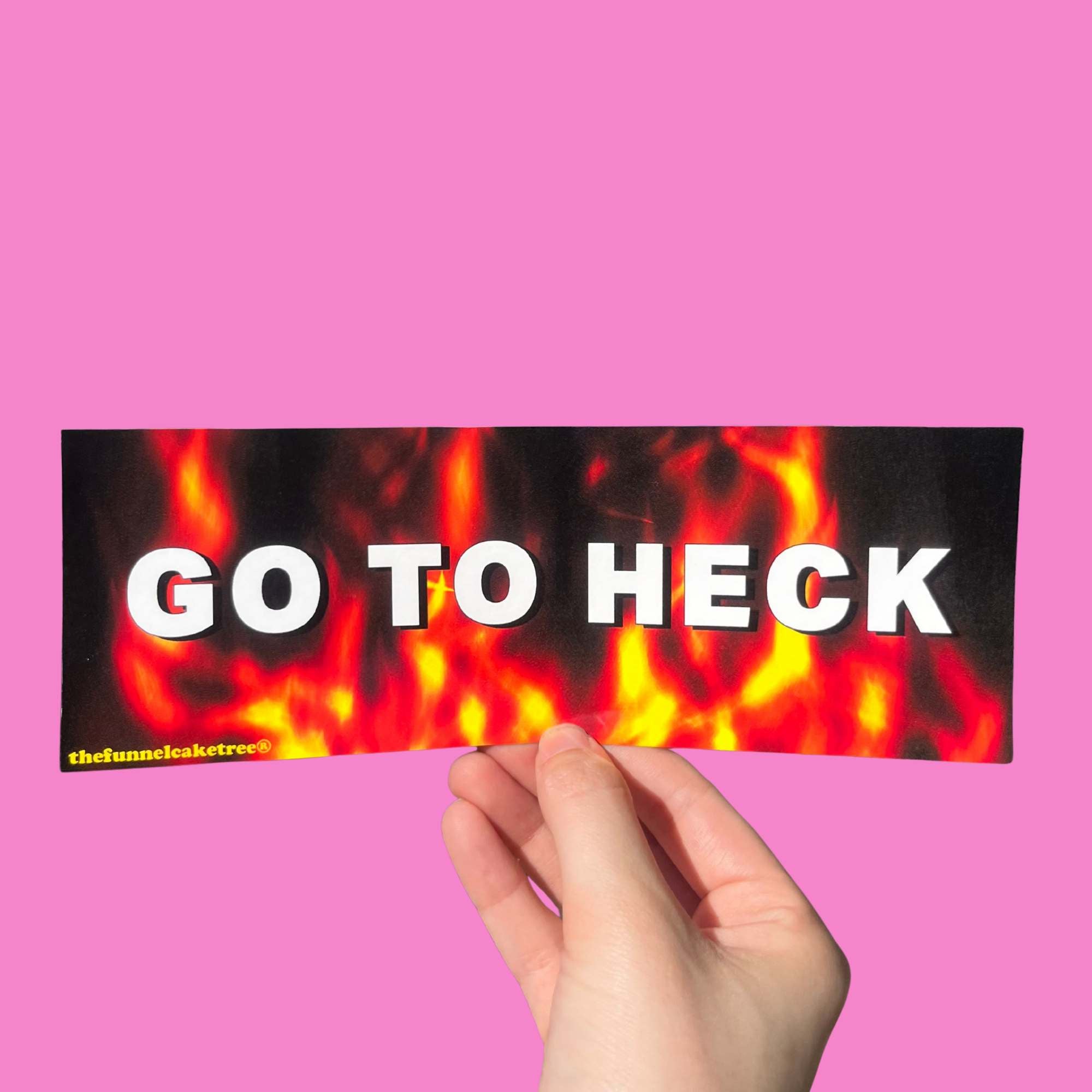 Glossy black bumper sticker with fiery red and yellow flames and bold white text reading “GO TO HECK,” held against a pink background.