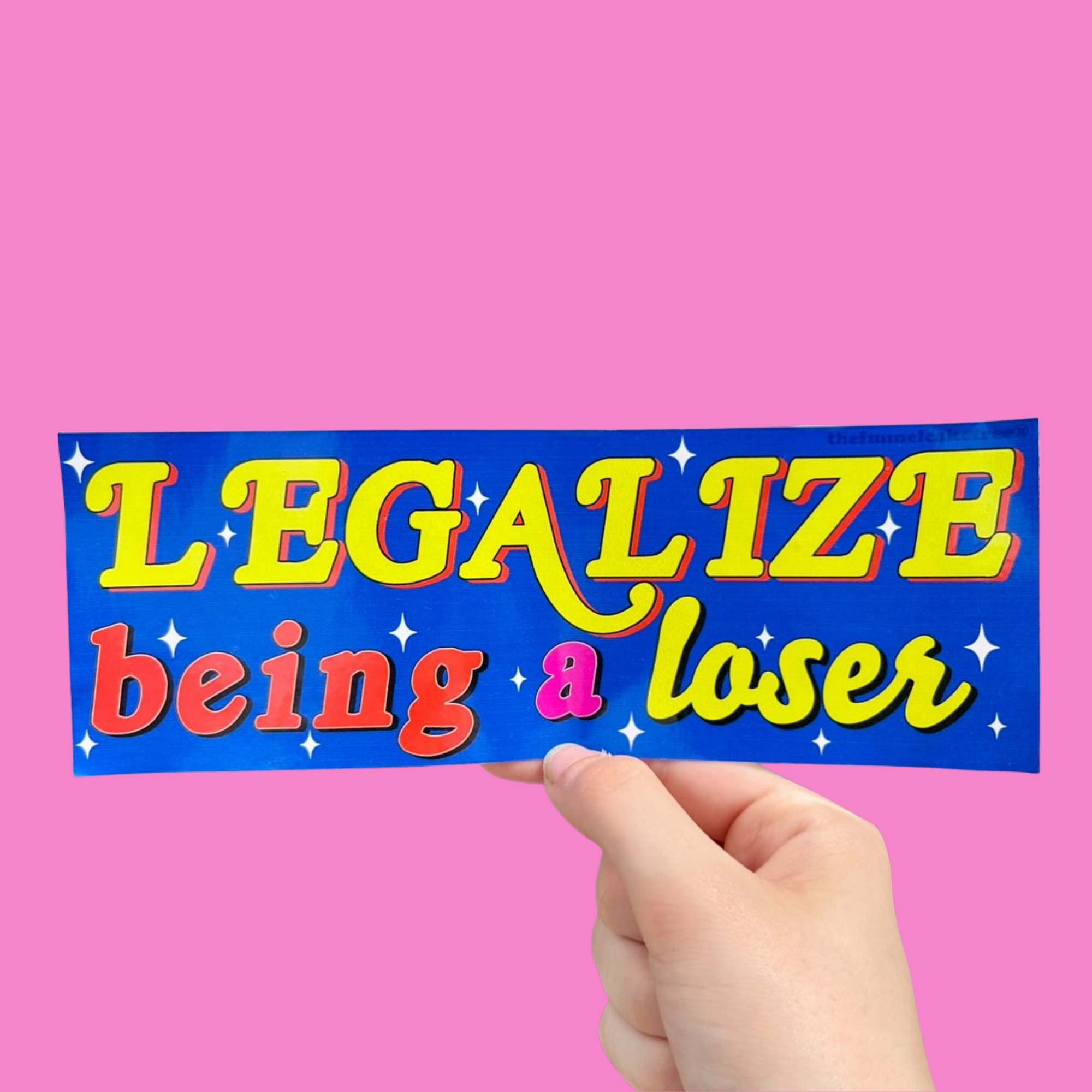 Glossy blue bumper sticker with yellow, red, and pink text reading “Legalize being a loser,” surrounded by white sparkles.
