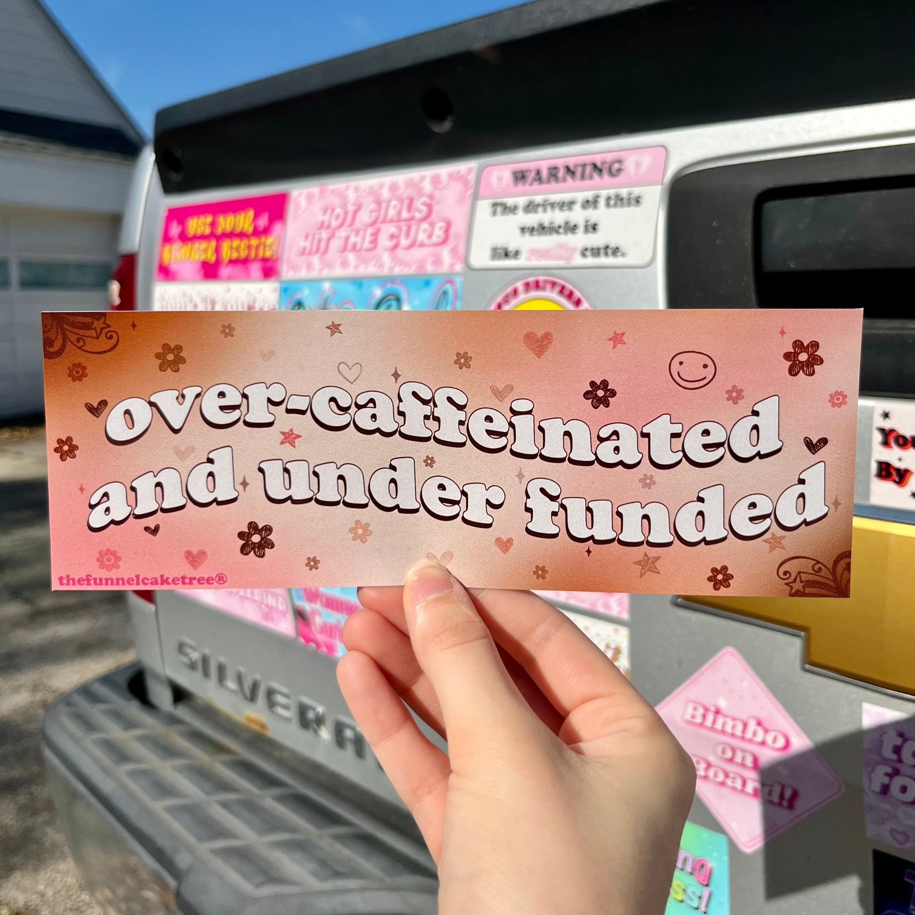 A hand holding a bumper sticker with the text 'overcaffineated and underfunded' on it, with a background featuring various other car stickers.