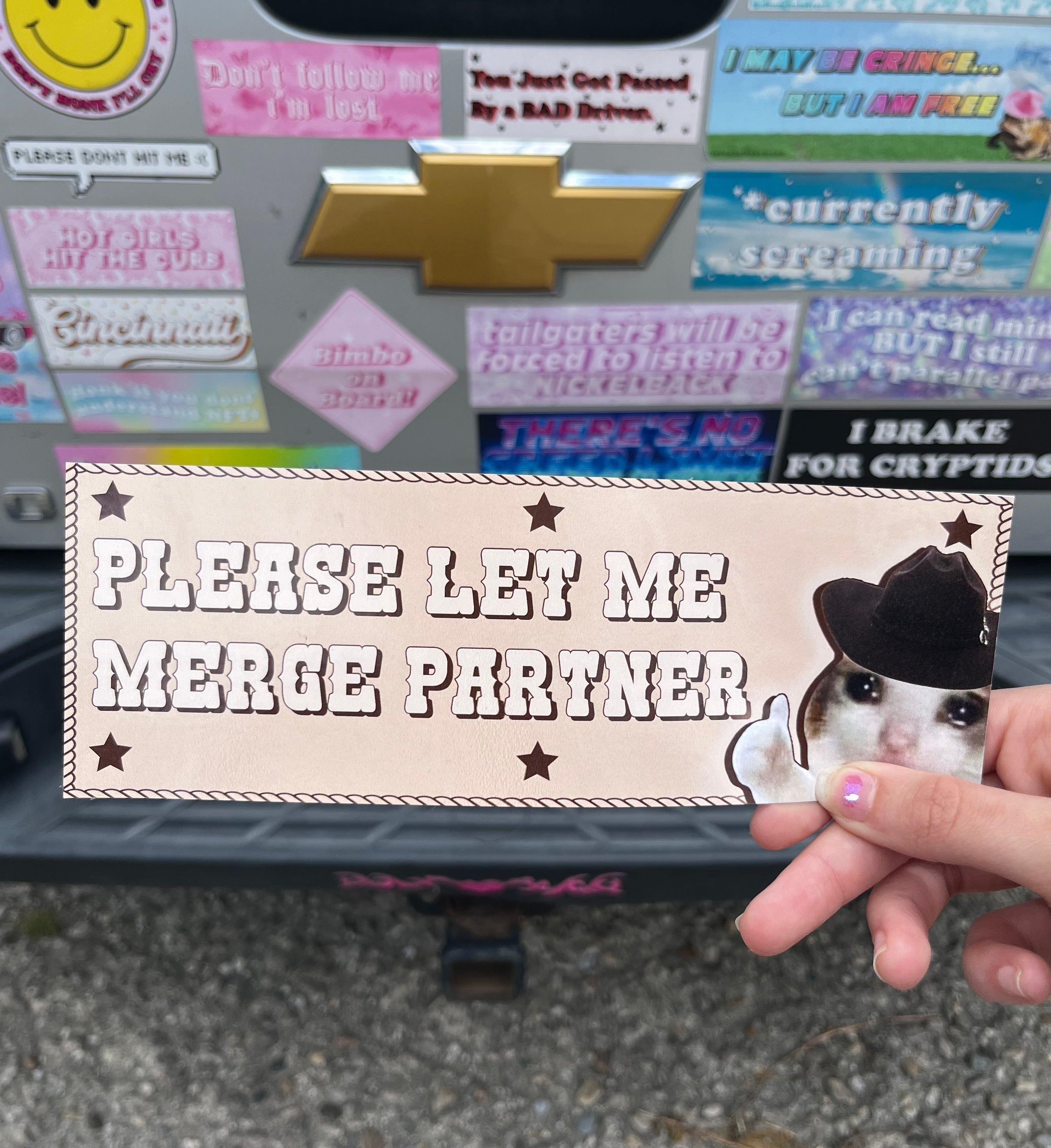 A hand holding a bumper sticker with text and a cartoon cat design that says 'Please let me merge partner' 
