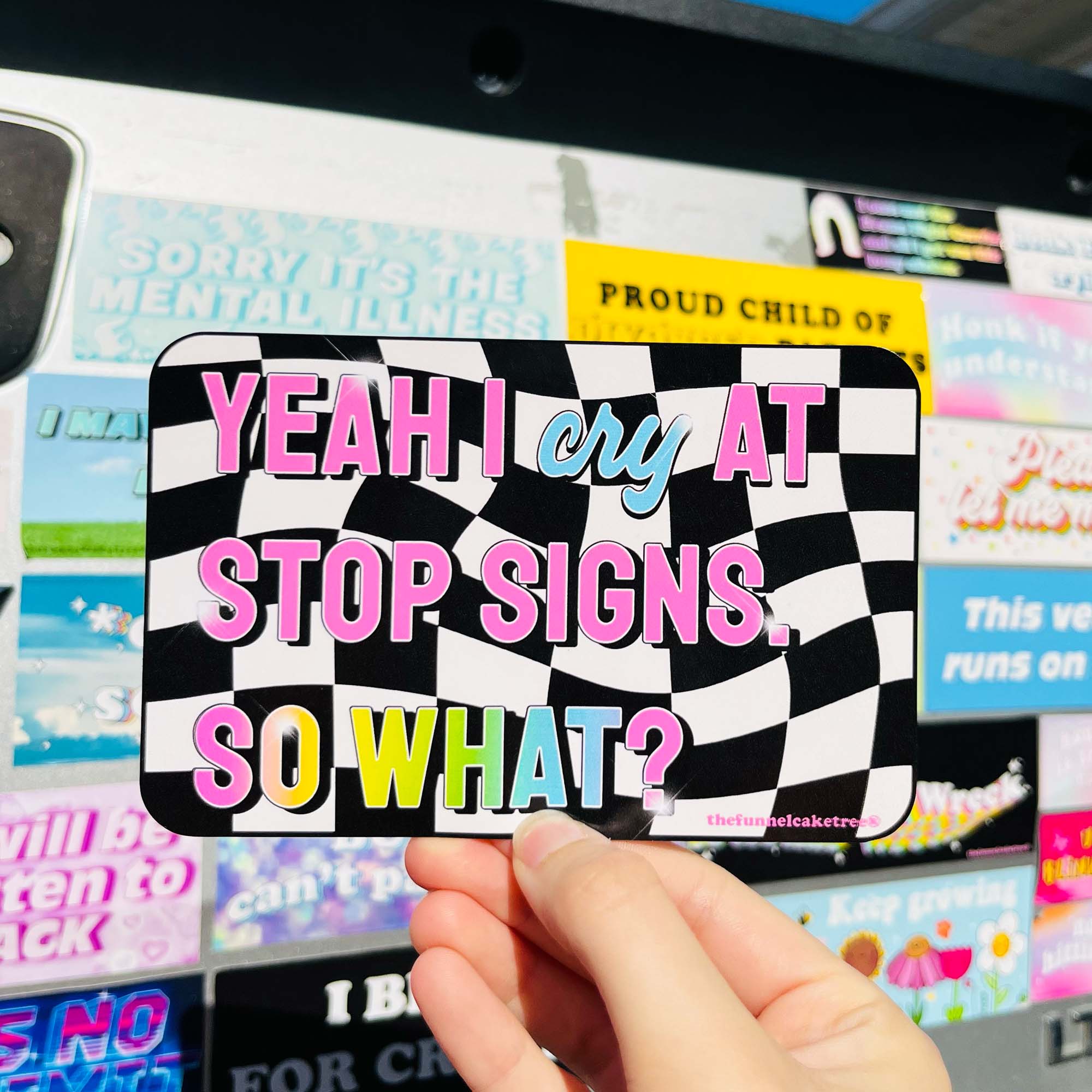 A hand holding a bumper sticker with a checkered black and white background and the text 'YEAH I cry at stop signs. SO WHAT?' in pink and other colors.