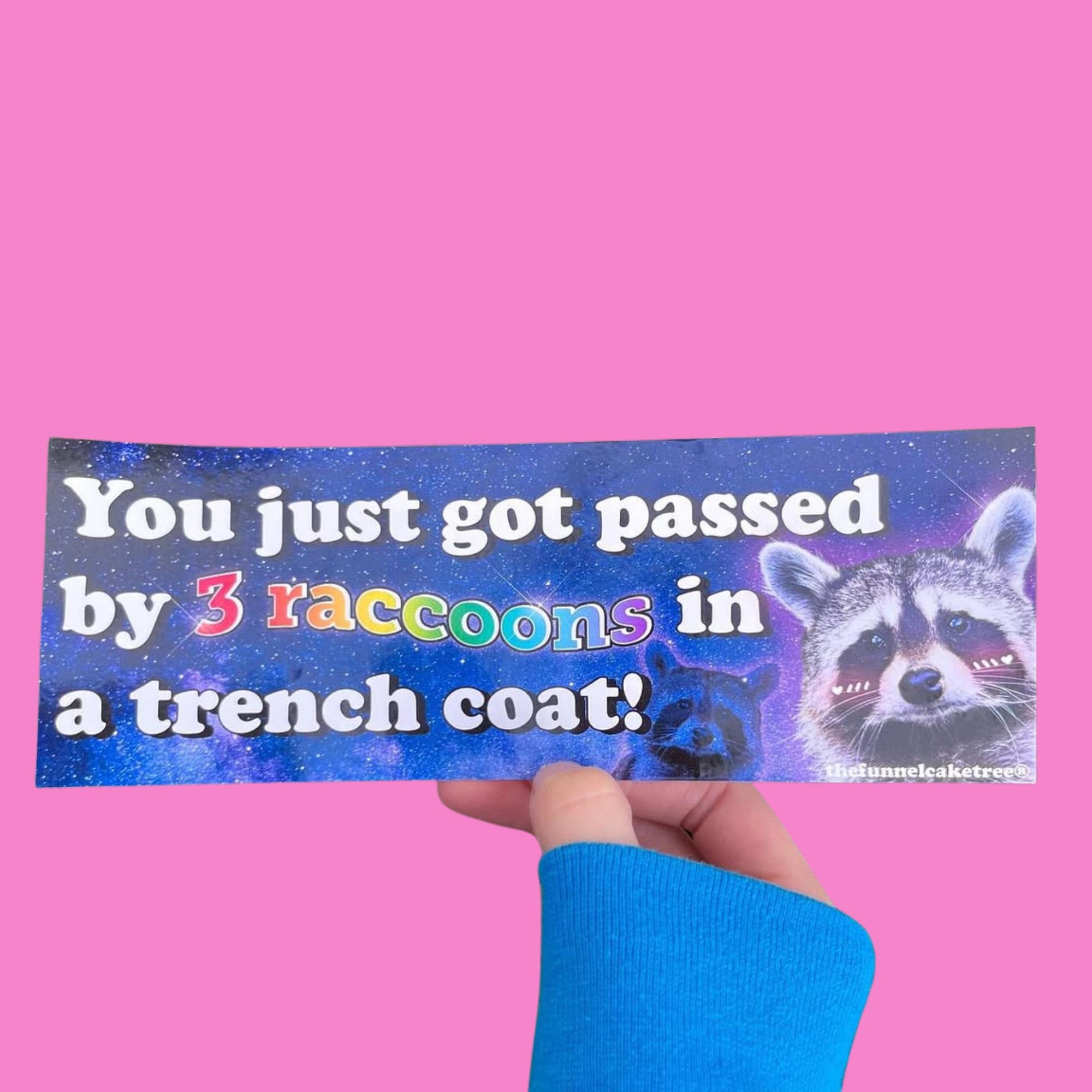 Glossy space-themed bumper sticker with white text reading “You just got passed by 3 raccoons in a trench coat!” featuring colorful lettering and illustrated raccoons with blushing cheeks on a cosmic background.