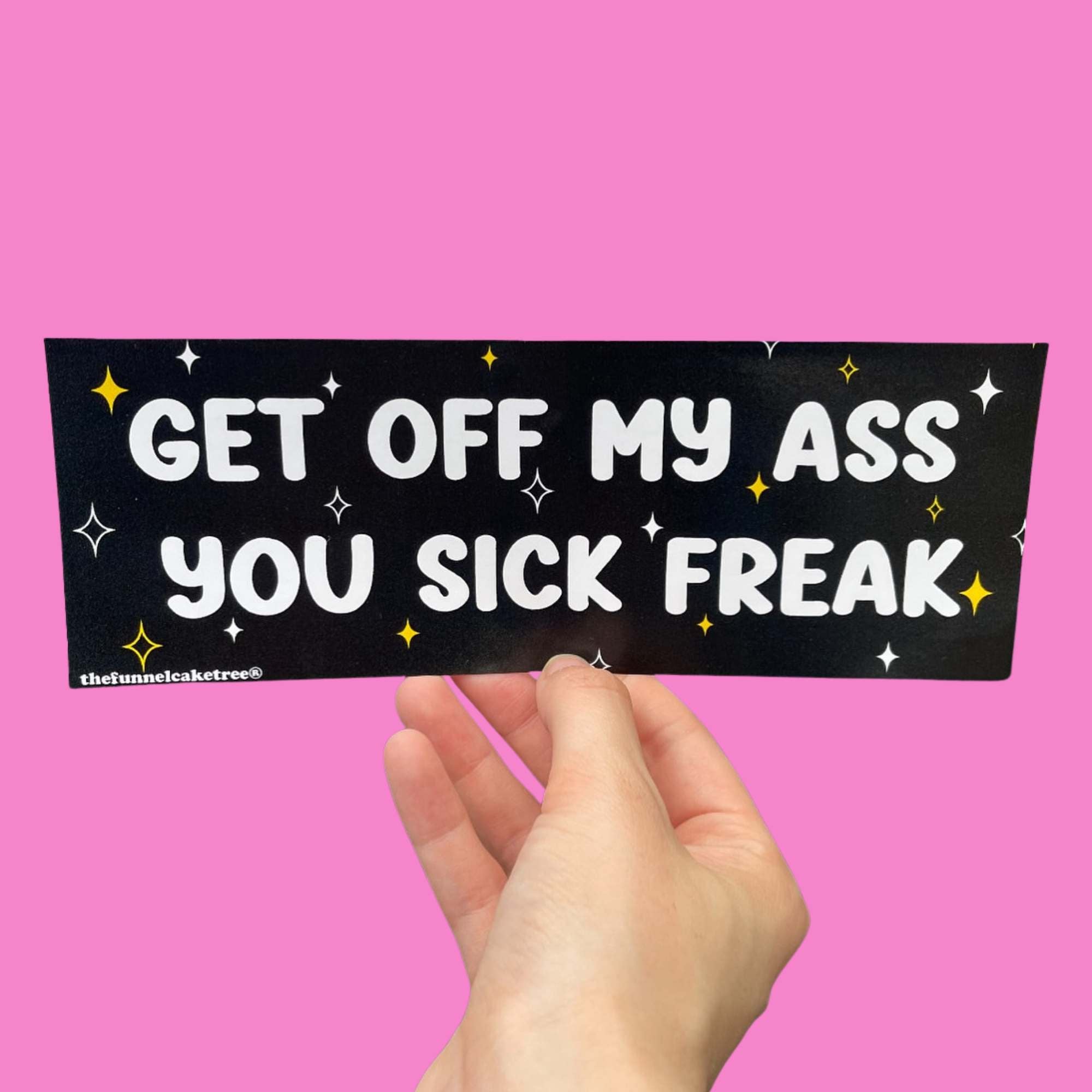 Glossy black bumper sticker with bold white text reading “GET OFF MY ASS YOU SICK FREAK,” surrounded by white and yellow sparkles, held against a pink background.