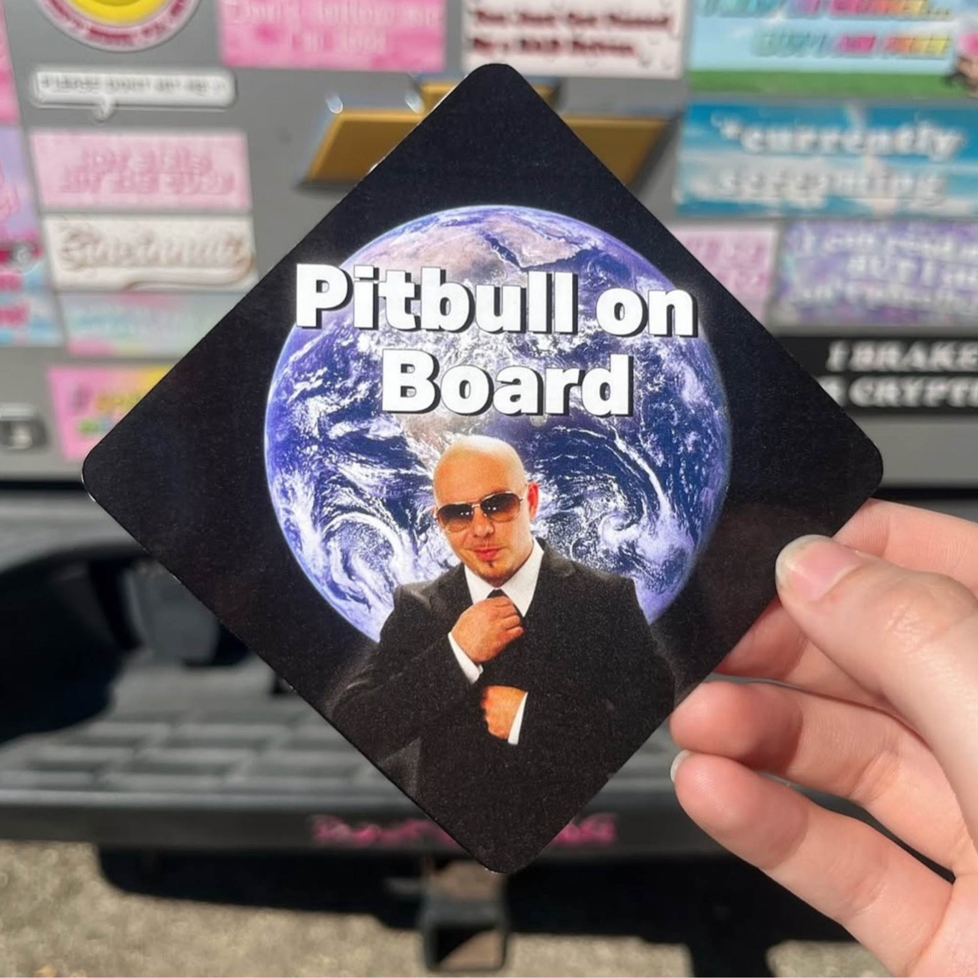 Sticker with a person in a suit and sunglasses and 'Pitbull on Board' text, held in front of a truck with stickers on the back. “Funny vinyl bumper sticker-Waterproof and UV-protected, perfect for cars.
