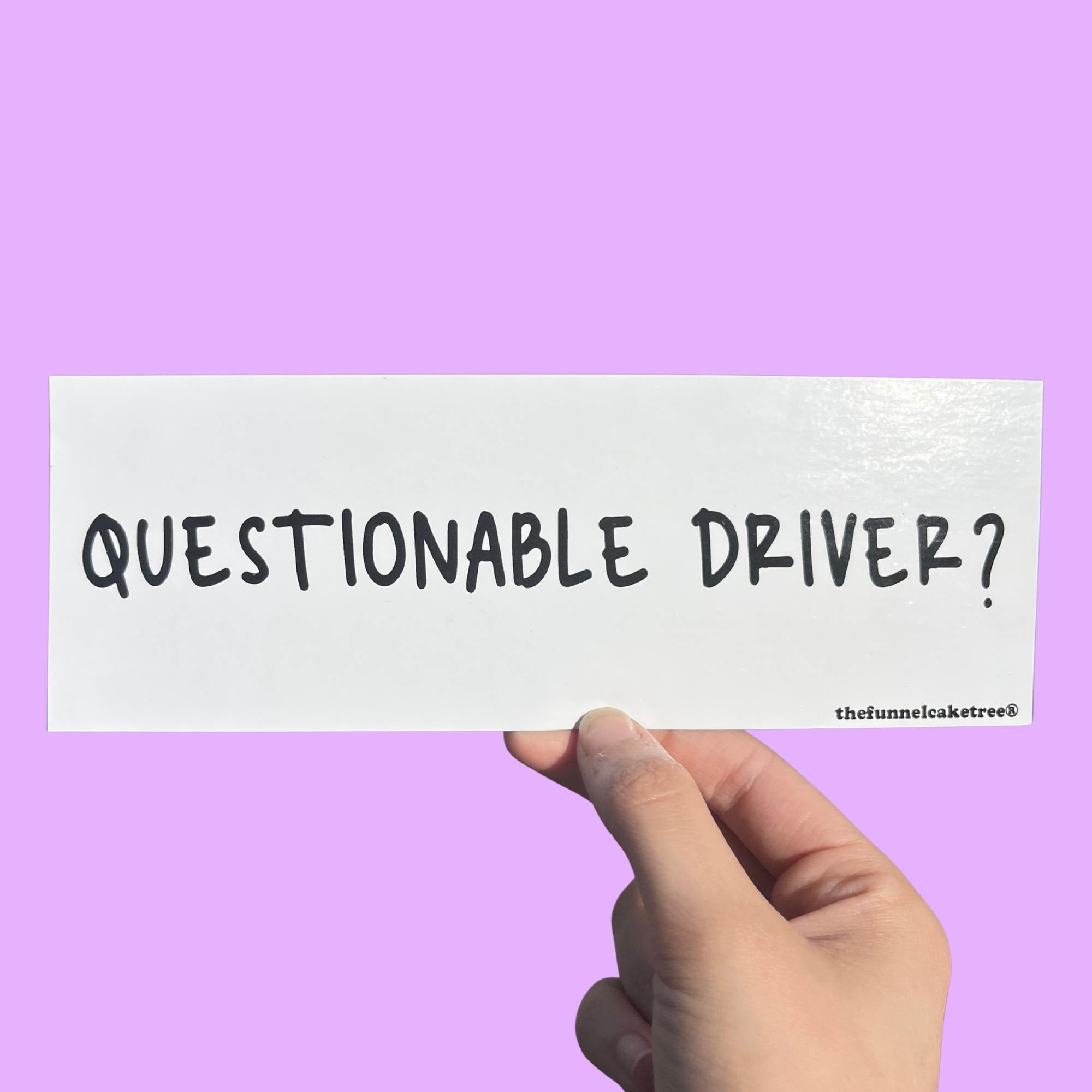 Glossy white bumper sticker with black text reading “QUESTIONABLE DRIVER?” held against a lavender background.