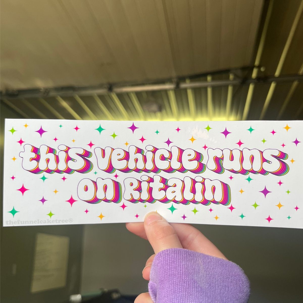 A hand holding a colorful bumper sticker with the text 'This Vehicle Runs on Ritalin' and multicolored stars and dots pattern.