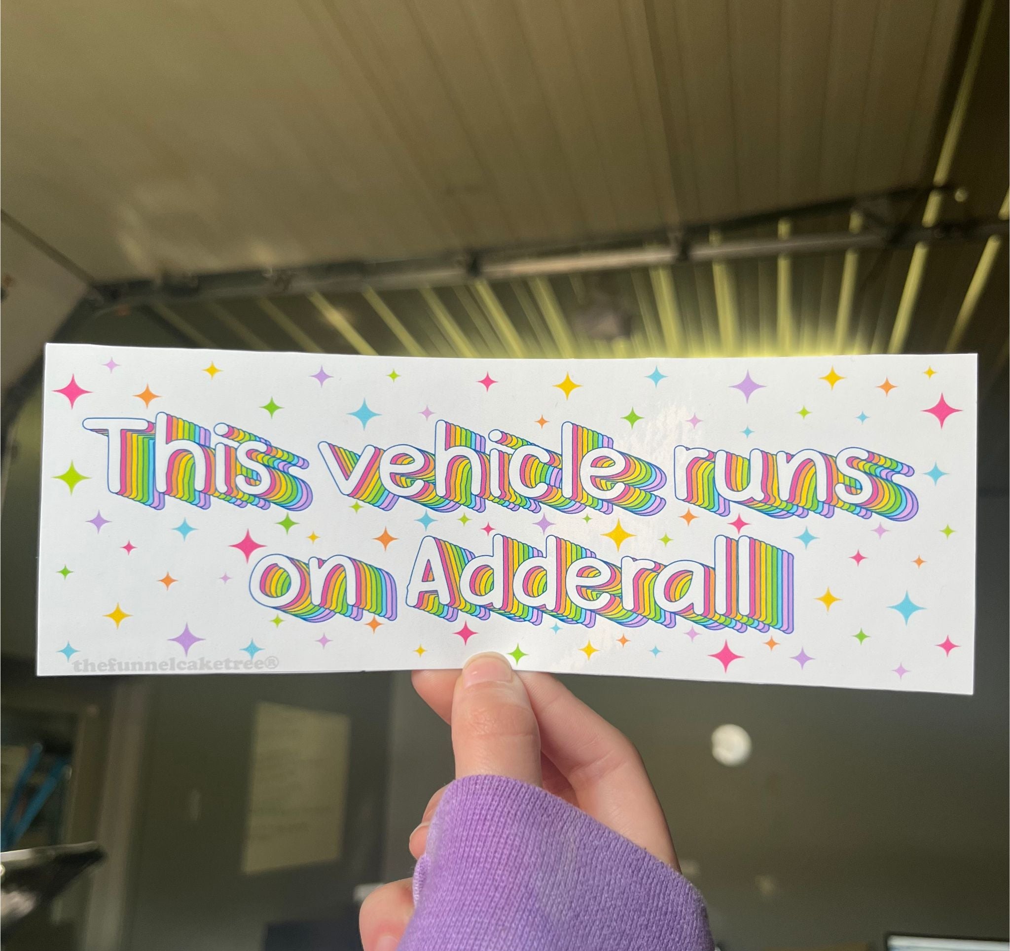 A hand holding a bumper sticker with the text 'This vehicle runs on Adderall' surrounded by colorful star graphics.