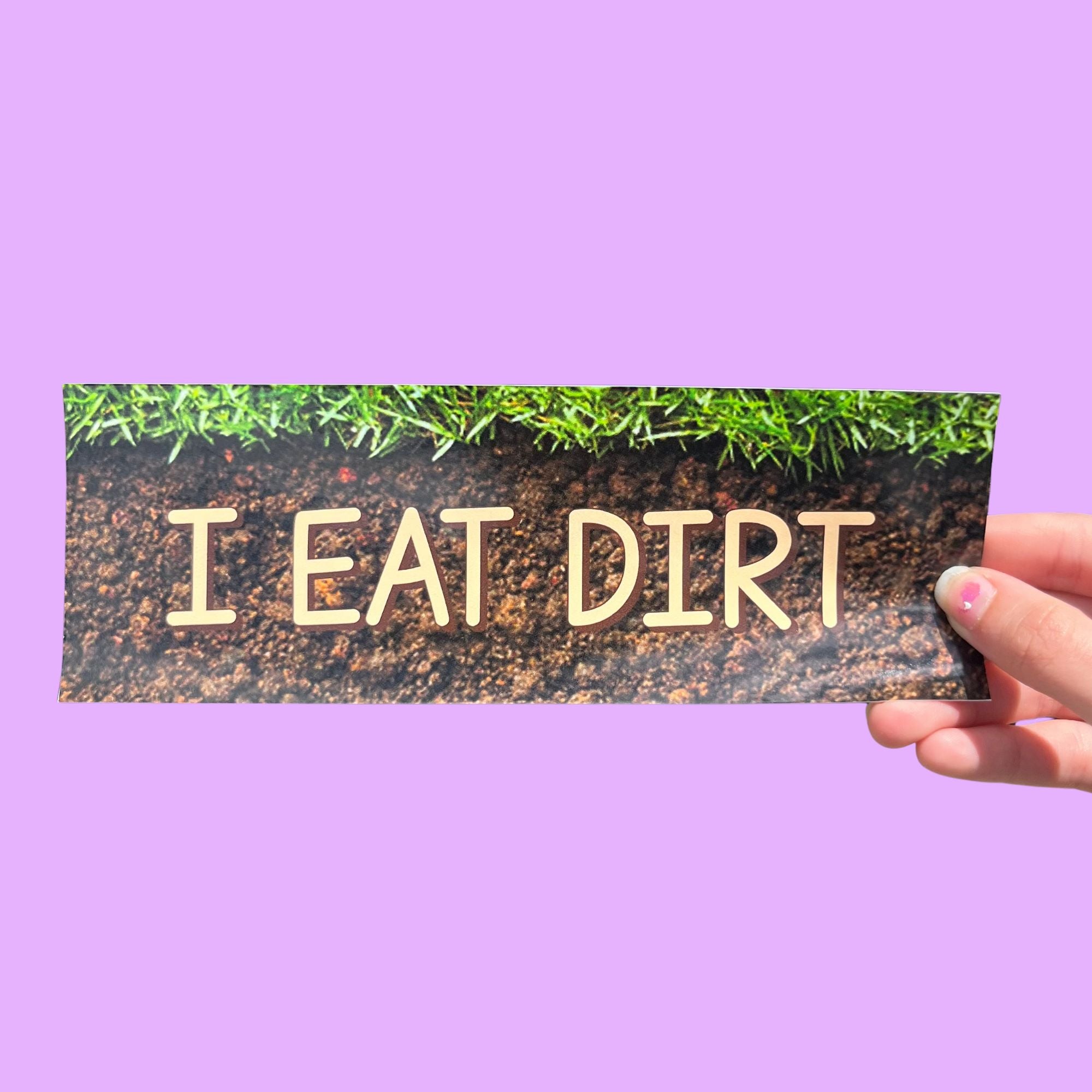 Glossy bumper sticker featuring soil and grass background with beige text reading “I EAT DIRT.”
