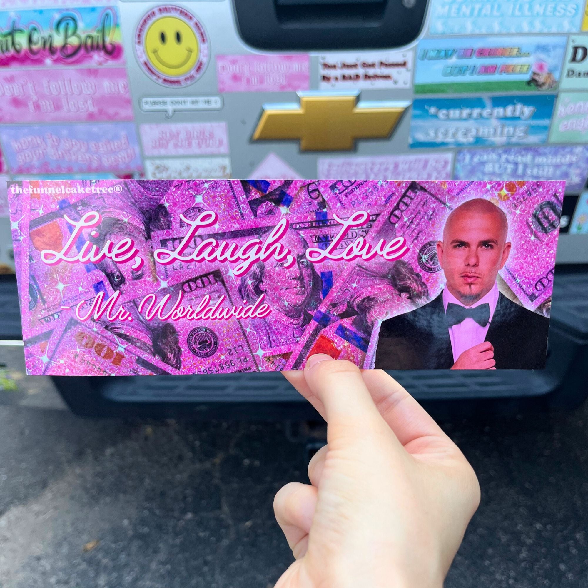 Hand holding a pink card with a person's face and text in front of a car with stickers.