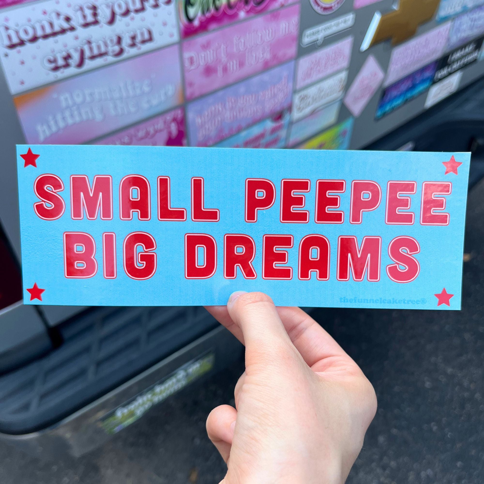 Hand holding a blue bumper sticker with red text 'SMALL PEEPEE BIG DREAMS' in front of the back of a truck bumper covered in various other bumper stickers.