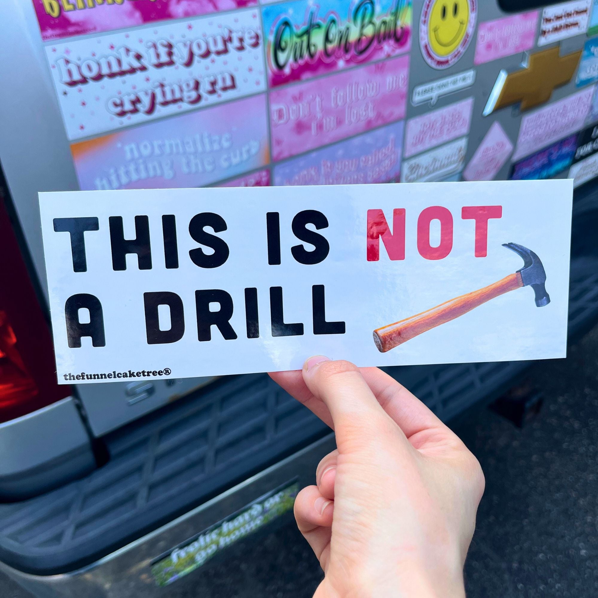 Hand holding a bumper sticker with text and a hammer graphic in front of a car with various stickers.