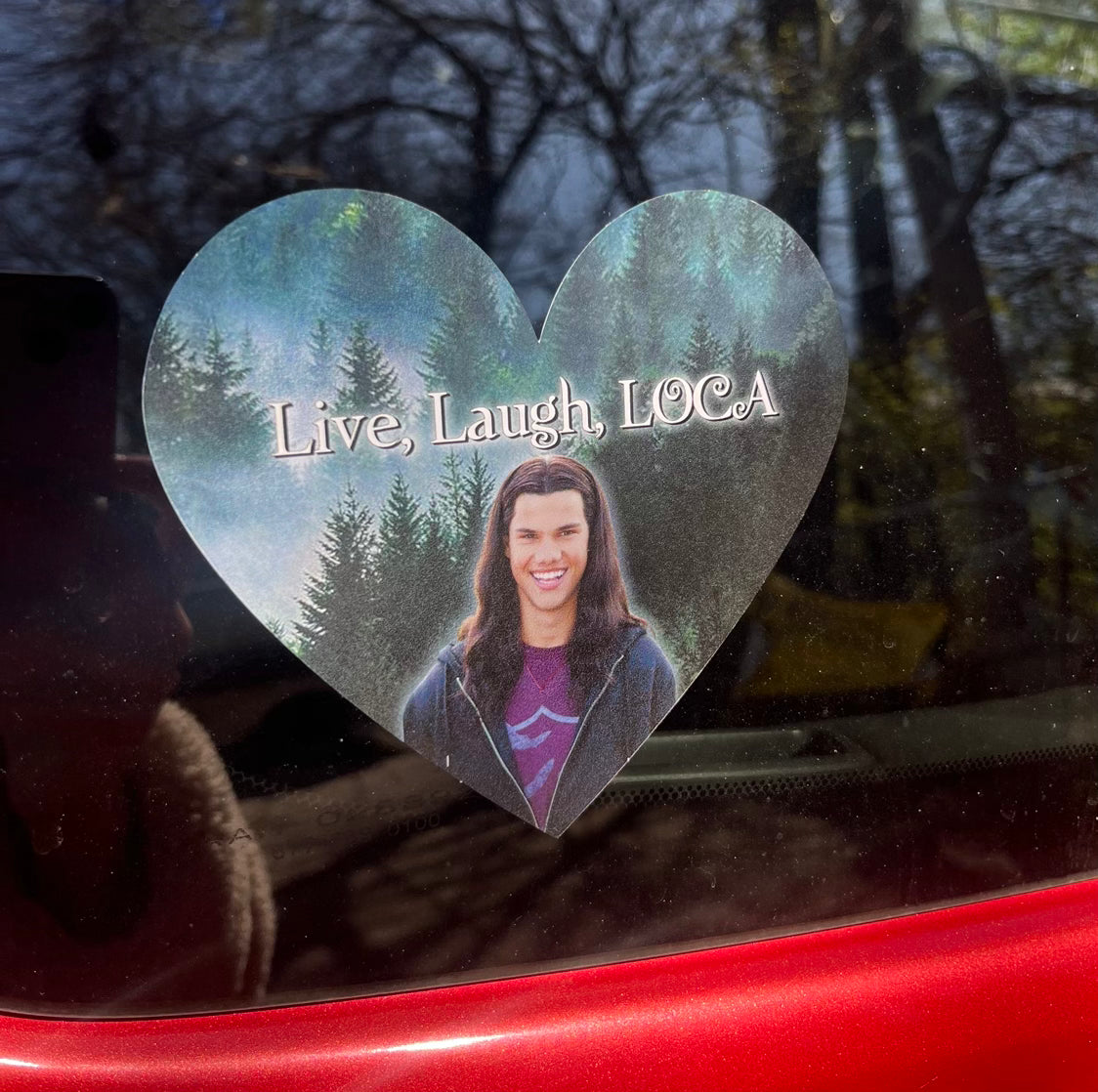 A heart-shaped bumper sticker with the text 'Live, Laugh, Loca' adhered to a red vehicle, featuring an image of a smiling person inside the heart shape.