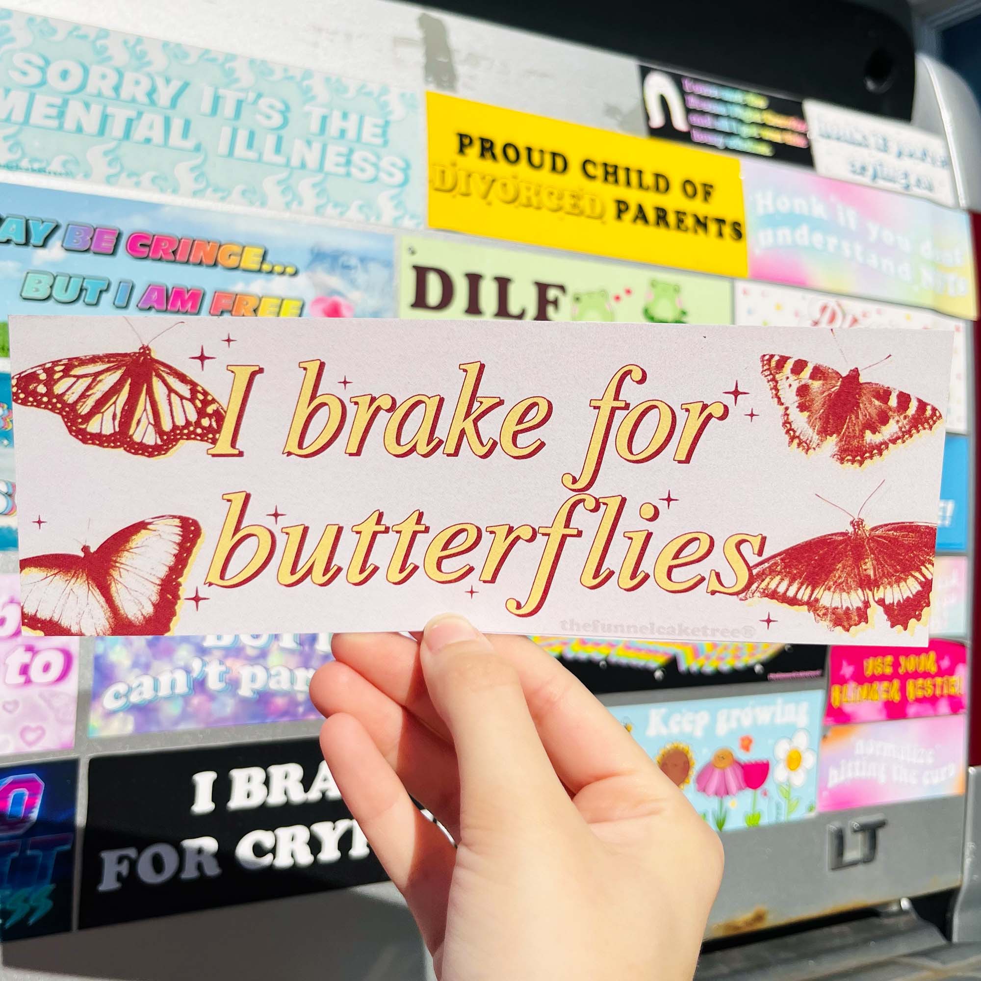 A hand holding a bumper sticker with the text 'I brake for butterflies' written on it, displayed against a background of various other car stickers.