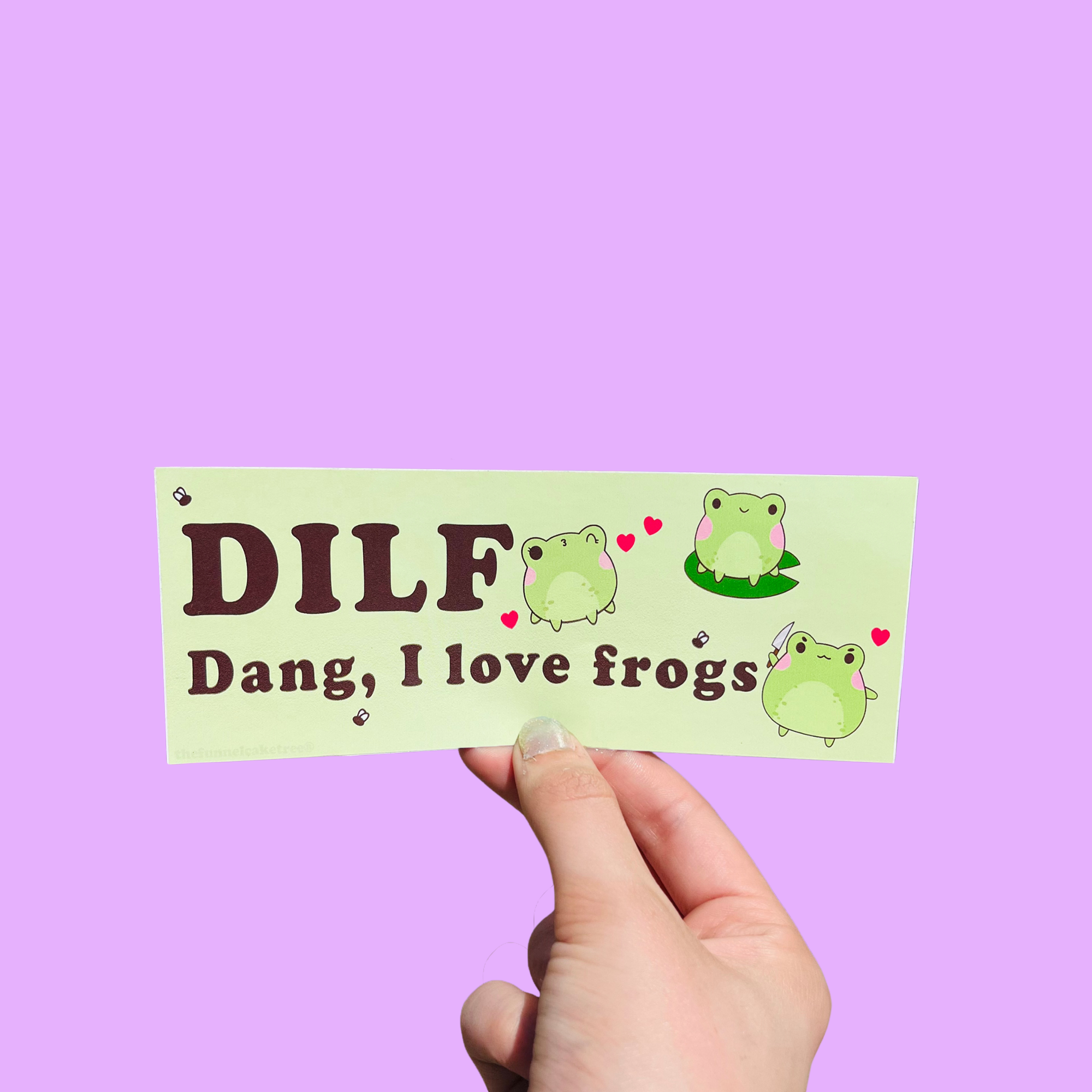Hand holding a green mini bumper sticker reading 'DILF — Dang, I love frogs,' with cartoon frogs and hearts On a solid purple background.