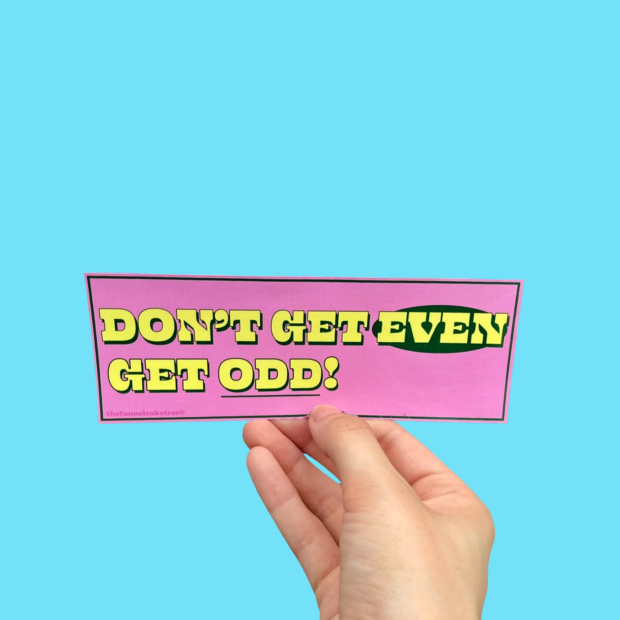 Hand holding a pink mini bumper sticker reading 'Don't get even! Get odd!' in yellow and green lettering On a solid teal background.