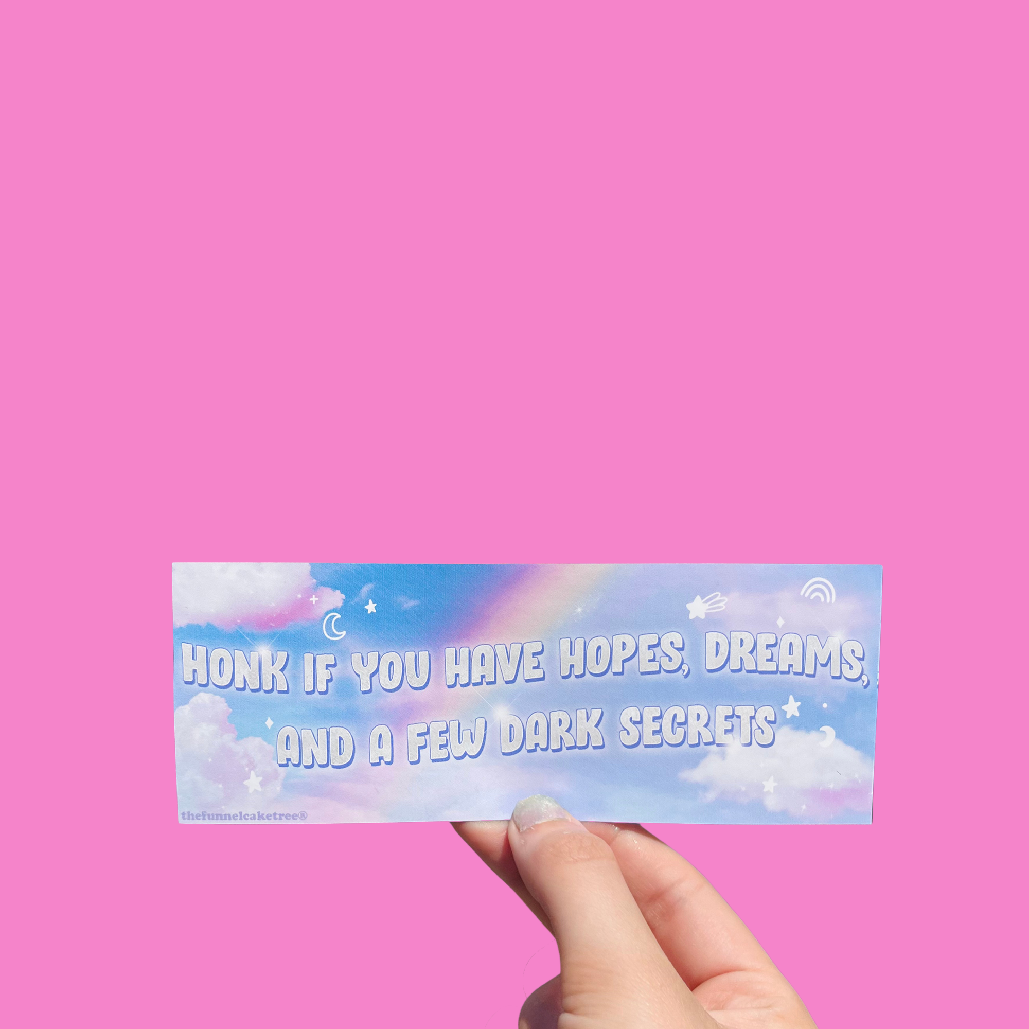 Hand holding a cloudy pastel mini sticker reading 'Honk if you have hopes, dreams and a few dark secrets.' On a solid pink background.