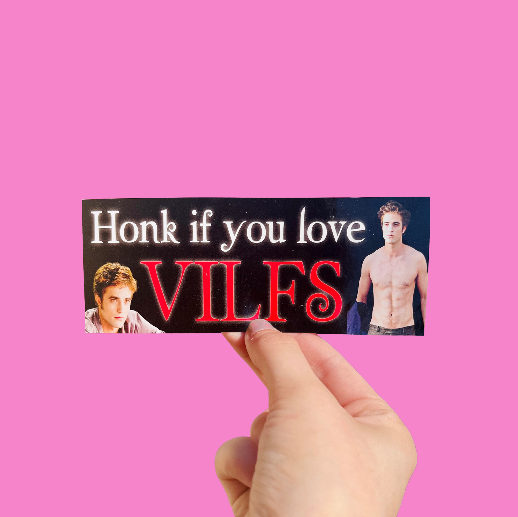 Hand holding a mini bumper sticker reading "honk if you love VILFS." On a solid pink background.