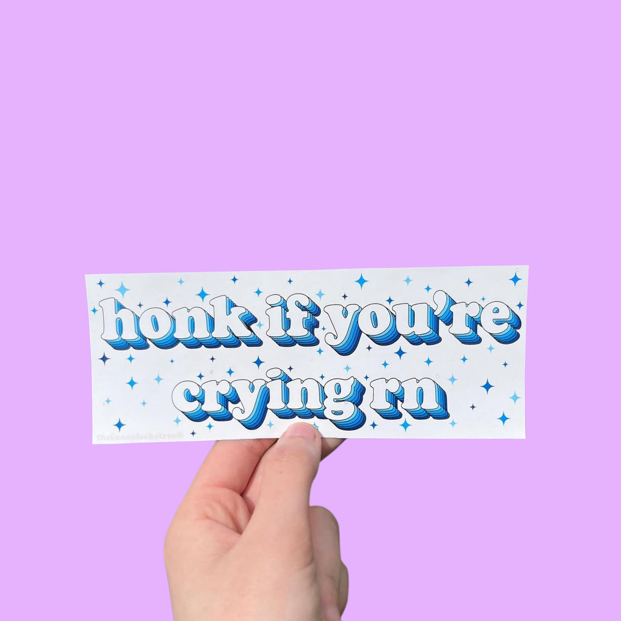 Hand holding a white mini bumper sticker reading 'honk if you're crying rn' in blue lettering with stars On a solid purple background.