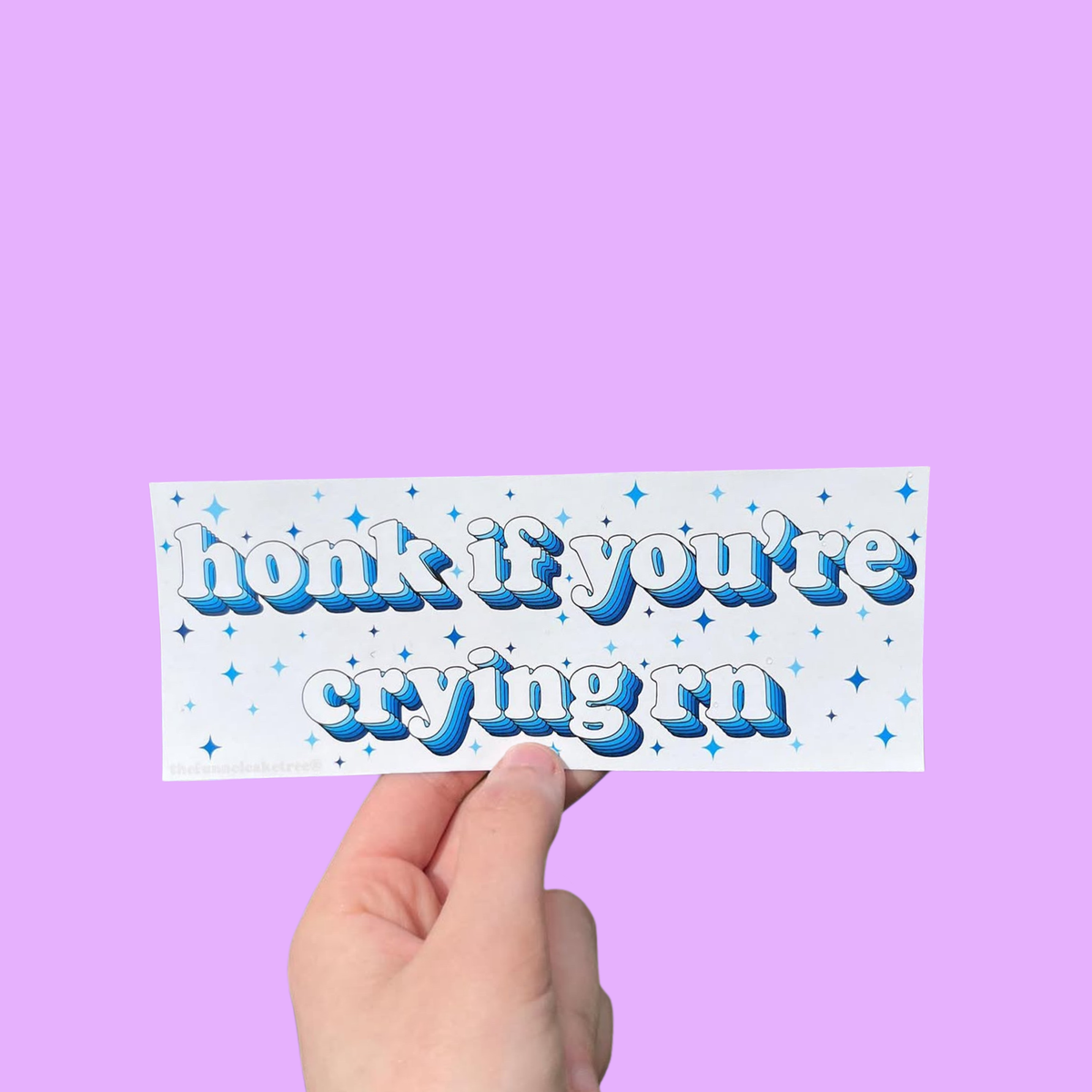 Hand holding a white mini bumper sticker reading 'honk if you're crying rn' in blue lettering with stars On a solid purple background.