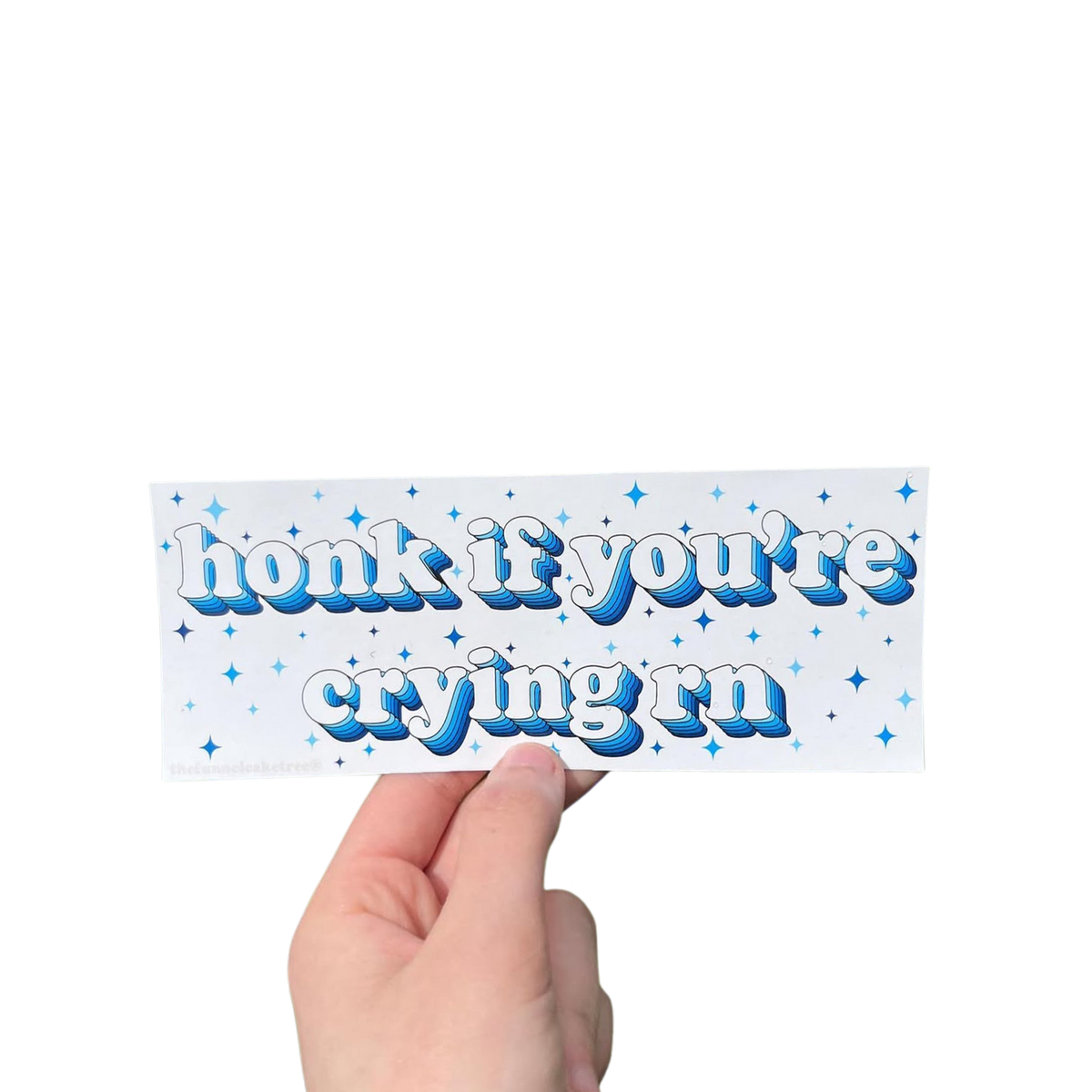 Hand holding a white mini bumper sticker reading 'honk if you're crying rn' in blue lettering with stars.