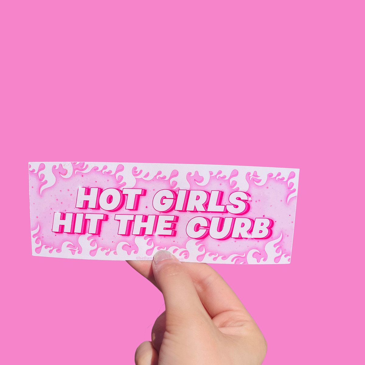 Hand holding a pink flame-patterned mini bumper sticker reading 'HOT GIRLS HIT THE CURB.' On a solid pink background.