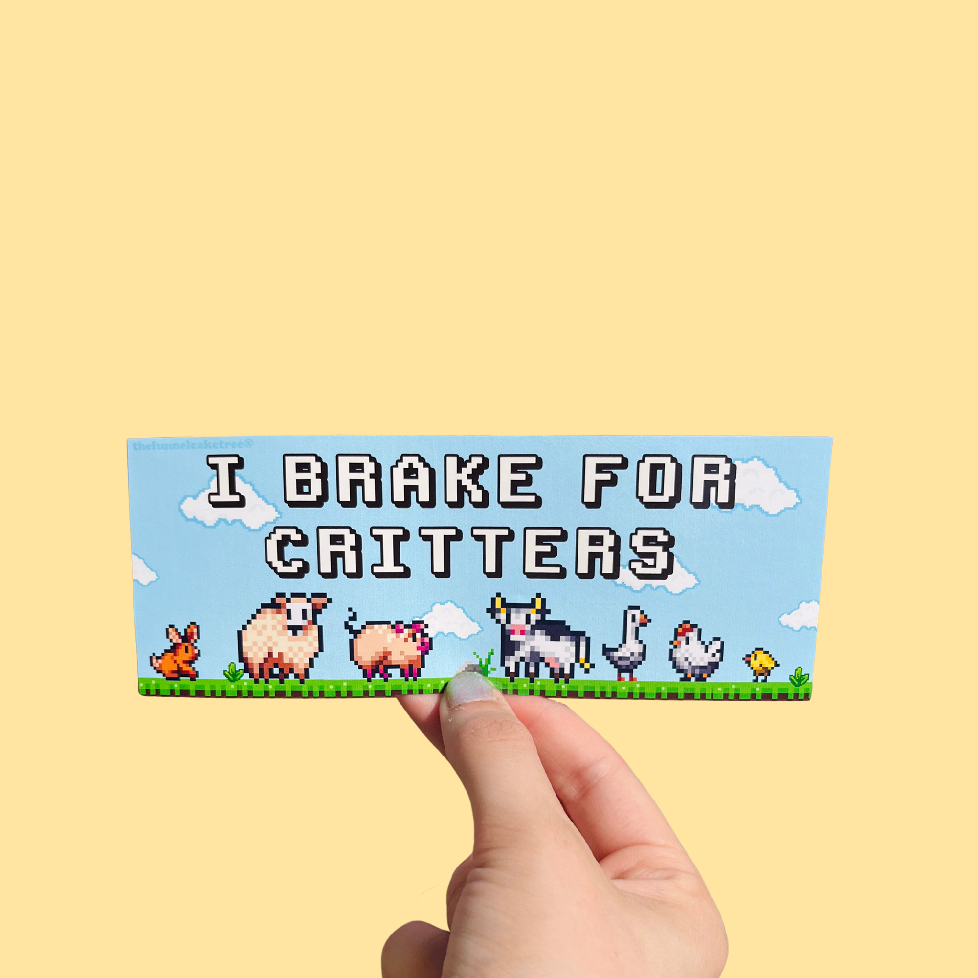 Hand holding a mini bumper sticker reading "i brake for critters." On a solid yellow background.