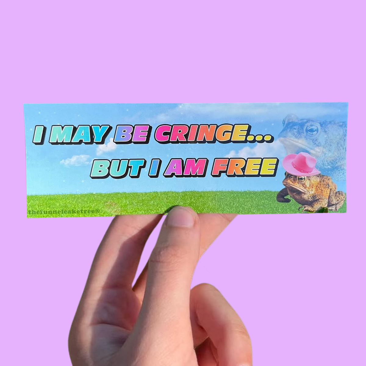 Hand holding a blue-sky mini sticker reading 'I may be cringe... but I am free,' with a frog blowing a pink bubble On a solid purple background.