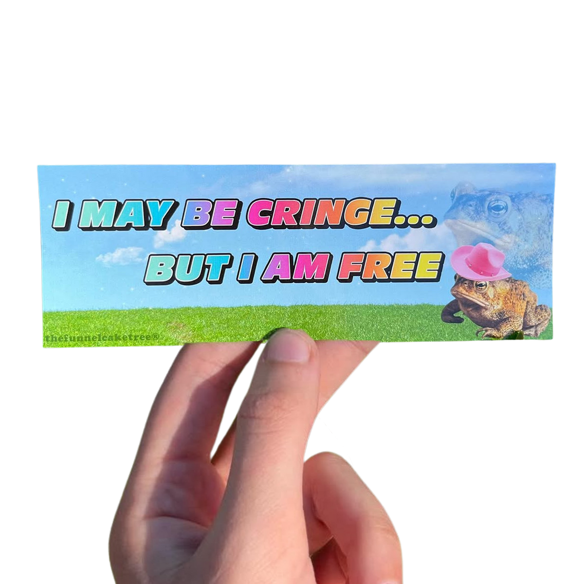 Hand holding a blue-sky mini sticker reading 'I may be cringe... but I am free,' with a frog blowing a pink bubble.