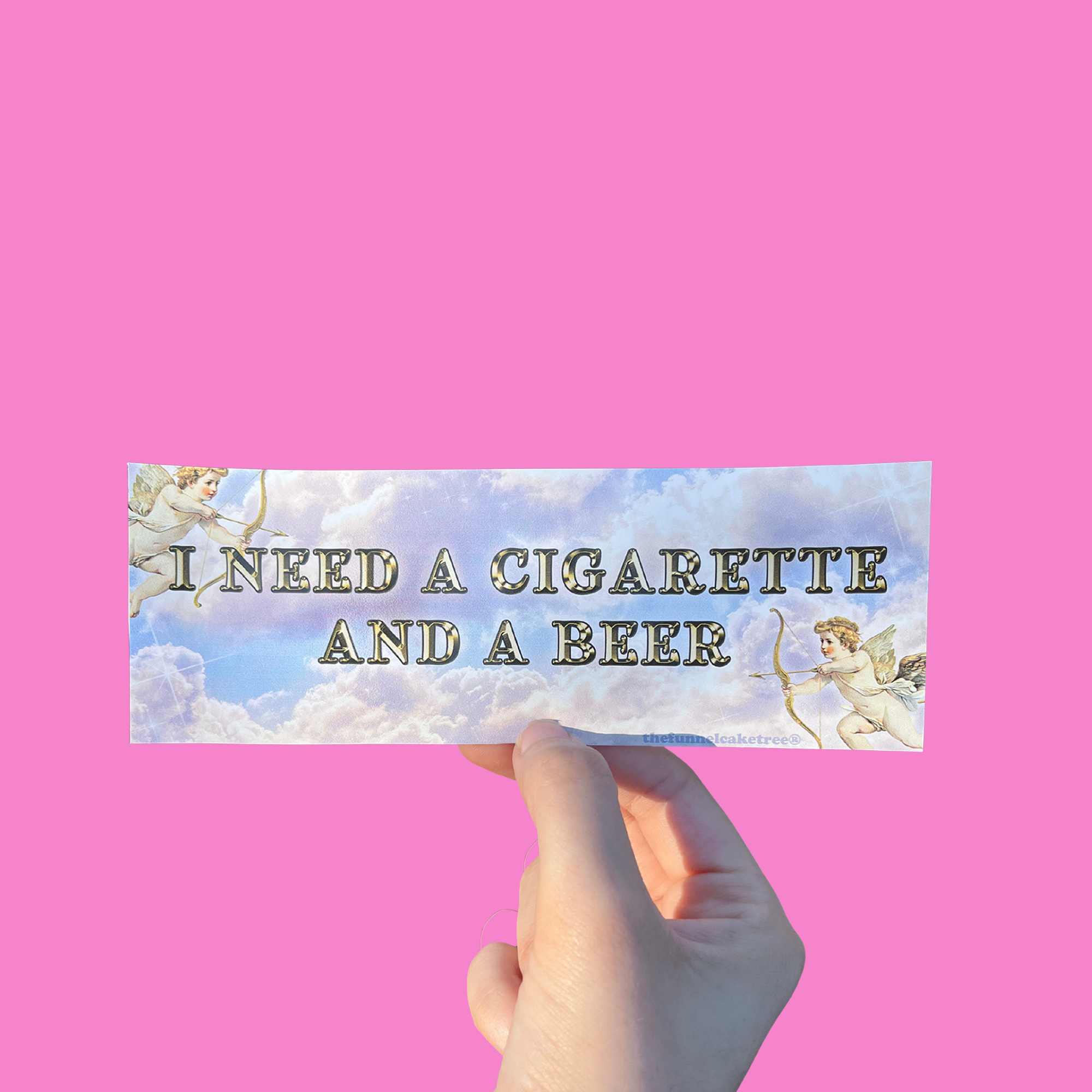 Hand holding a cloudy mini bumper sticker reading 'I NEED A CIGARETTE AND A BEER' beside a cherub illustration On a solid pink background.