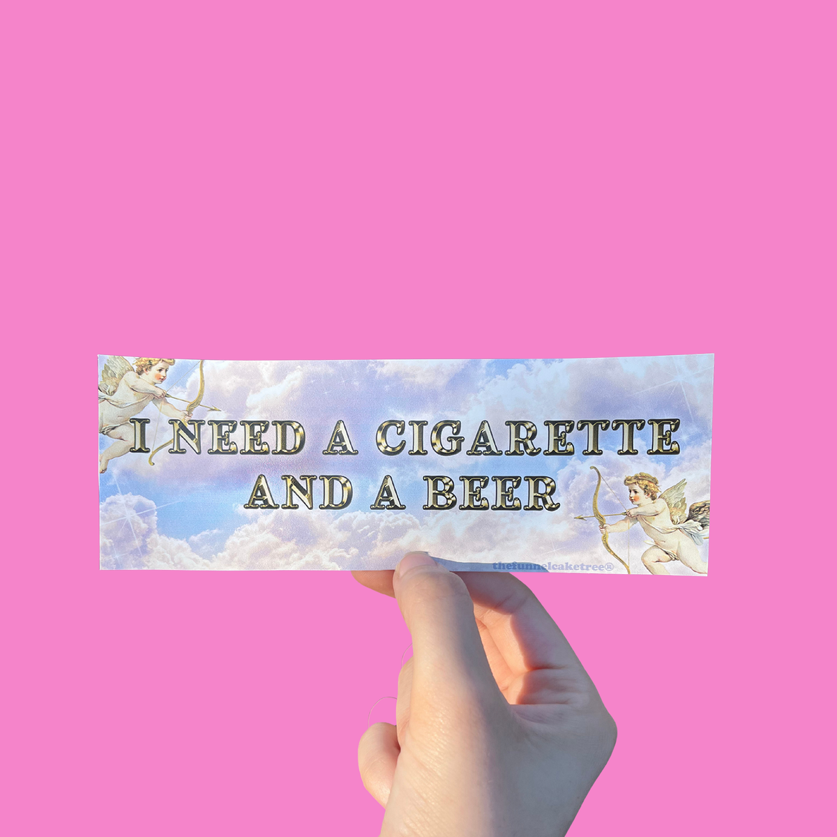 Hand holding a cloudy mini bumper sticker reading 'I NEED A CIGARETTE AND A BEER' beside a cherub illustration On a solid pink background.